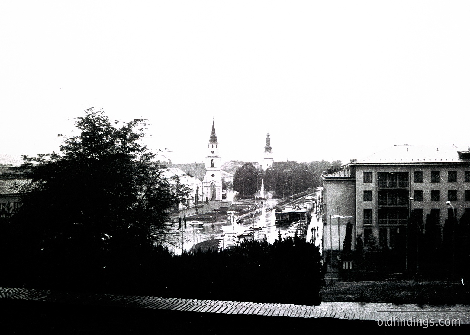 Early 20th-century urban scene featuring a church with a tall steeple and a monument in the center. Cobblestone streets and sparse traffic suggest a quiet, possibly pre-automotive era. Surrounding architecture includes multi-story buildings with classical facades. Likely Eastern European due to architectural style.