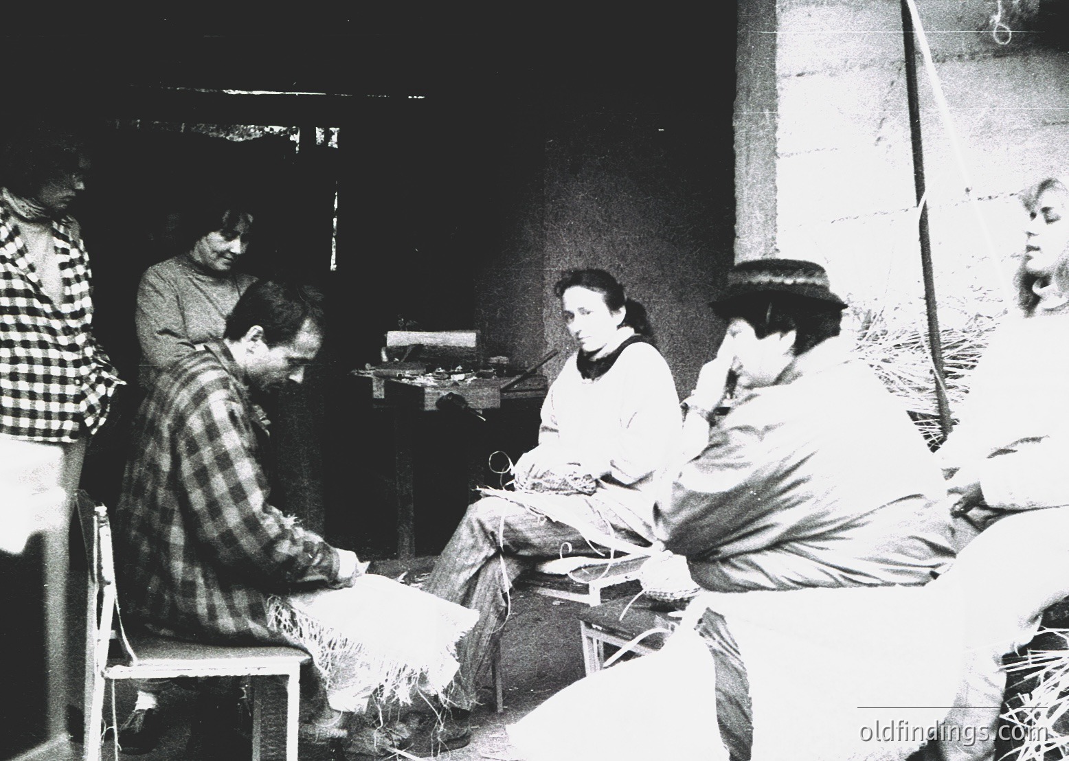 Group of five individuals engaged in textile work in a dimly lit, rustic indoor setting, likely a workshop or communal space. Central figure sits at a spinning wheel, surrounded by others holding yarn or fabric. Mid-century clothing and simple furnishings suggest a 1950s–1960s European or Middle Eastern rural scene.