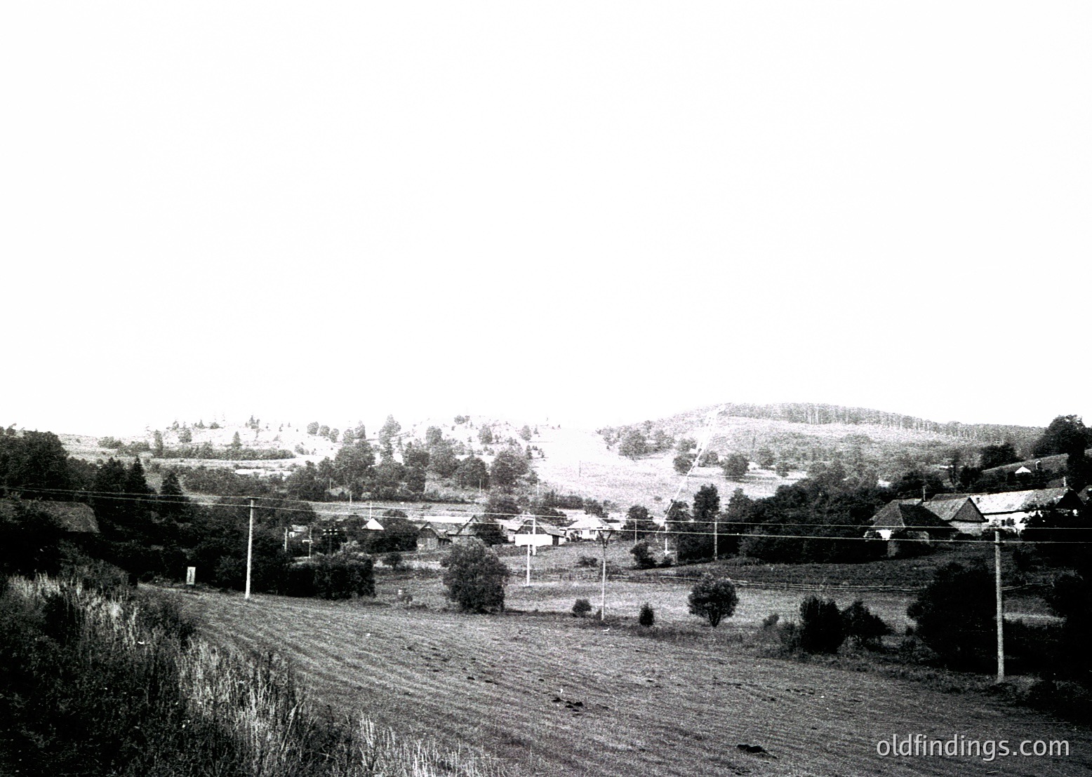Rural landscape featuring scattered farmhouses and dense forest patches, framed by power lines and utility poles. Mid-20th century agricultural setting with open fields and rolling terrain.