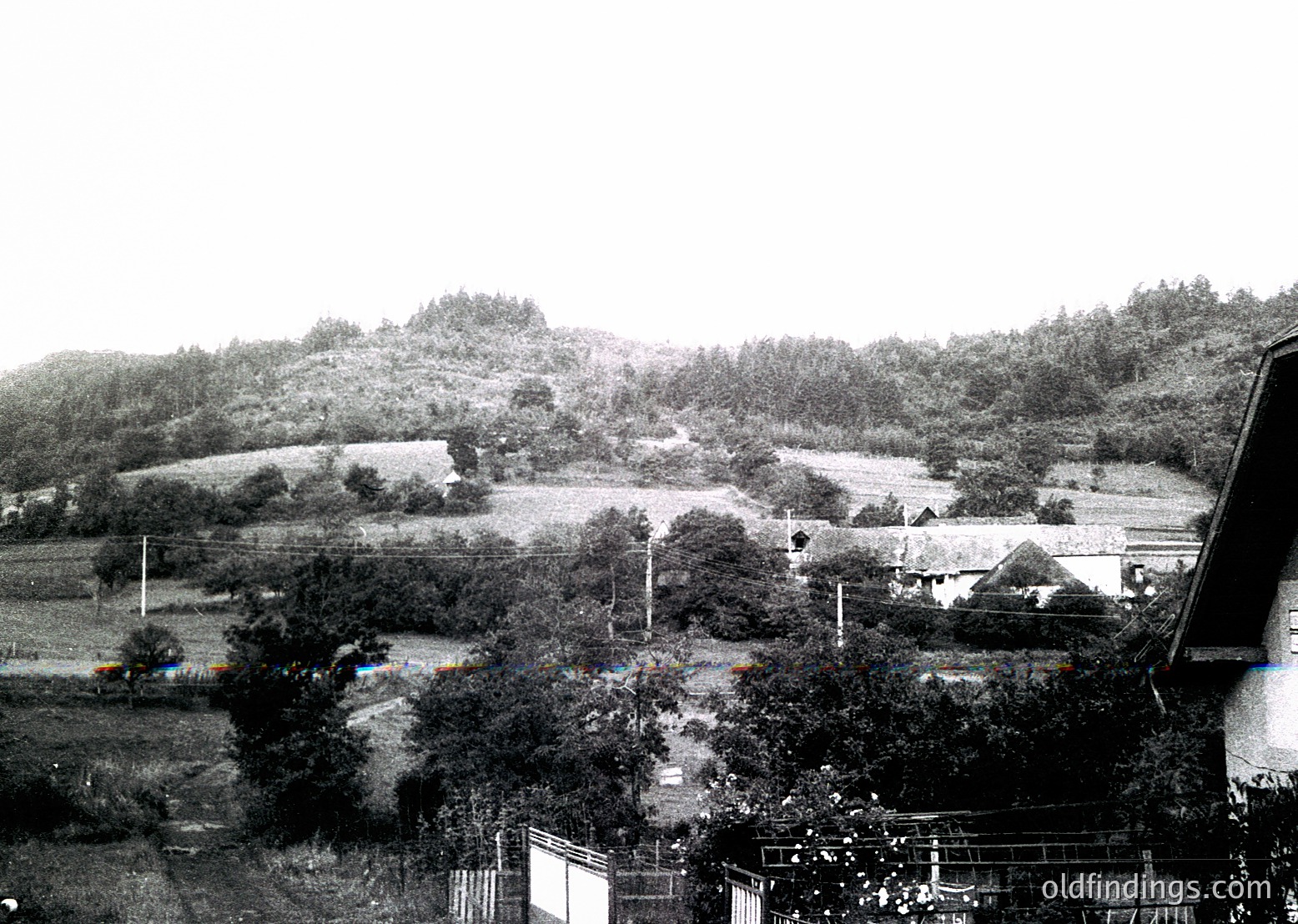Mid-20th century rural landscape featuring terraced farmland with scattered wooden structures. Overcast skies and dense foliage dominate the scene, suggesting agricultural activity in a hilly region. Fence and utility poles indicate infrastructure development.