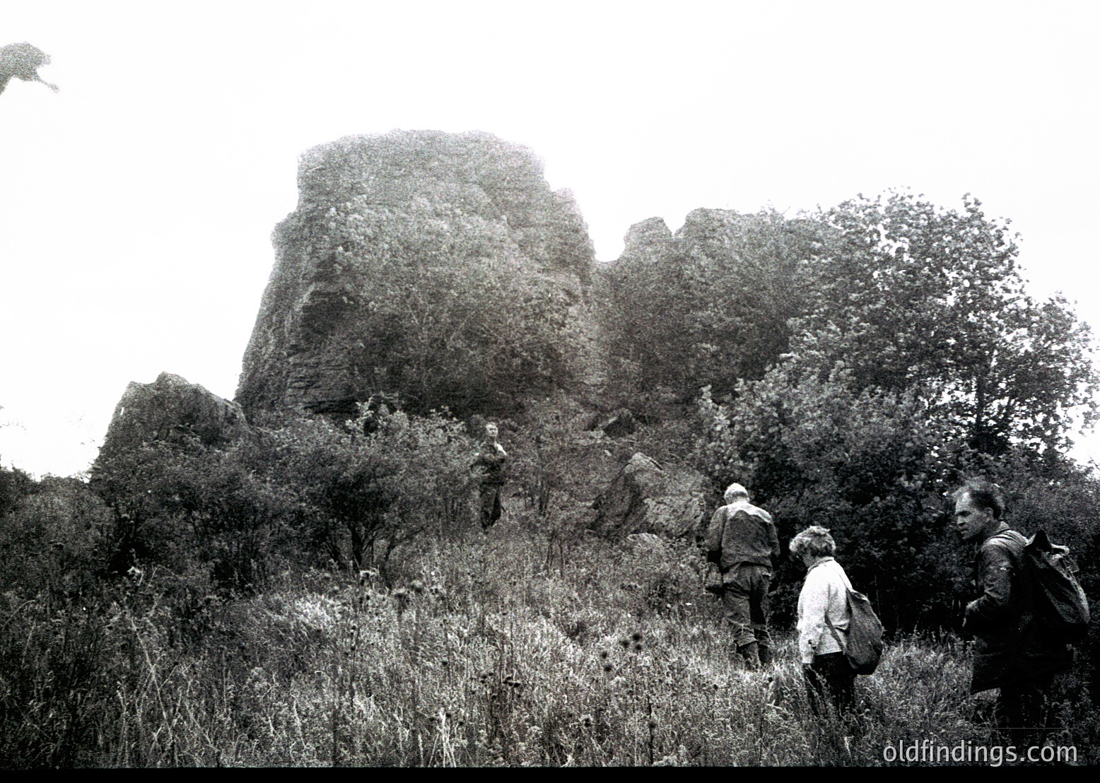 Black-and-white shot of hikers ascending a rugged rock formation, likely a historic fortress or natural landmark. Mid-20th century attire suggests or . Dense vegetation and steep terrain indicate a European or Mediterranean setting. Ideal for historical research or outdoor adventure themes.