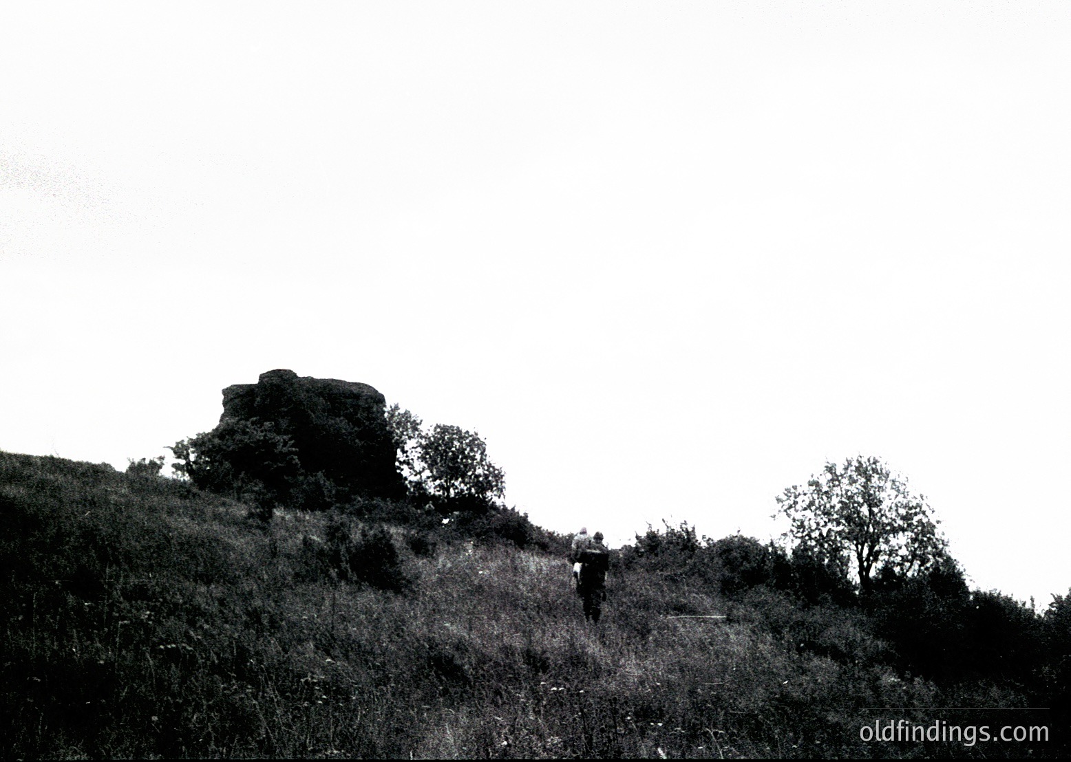 Black-and-white landscape featuring lone figure ascending a grassy hill toward a prominent rock formation. Mid-20th century outdoor photography style.