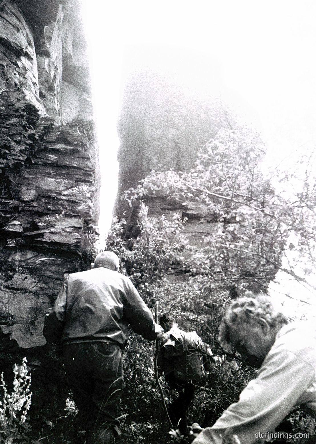 Two men engage in rock climbing in rugged terrain, mid-19th to early 20th century. One climber ascends a steep, moss-covered rock face while the other secures him with a rope. The scene captures early mountaineering techniques and the natural landscape’s vertical challenges.