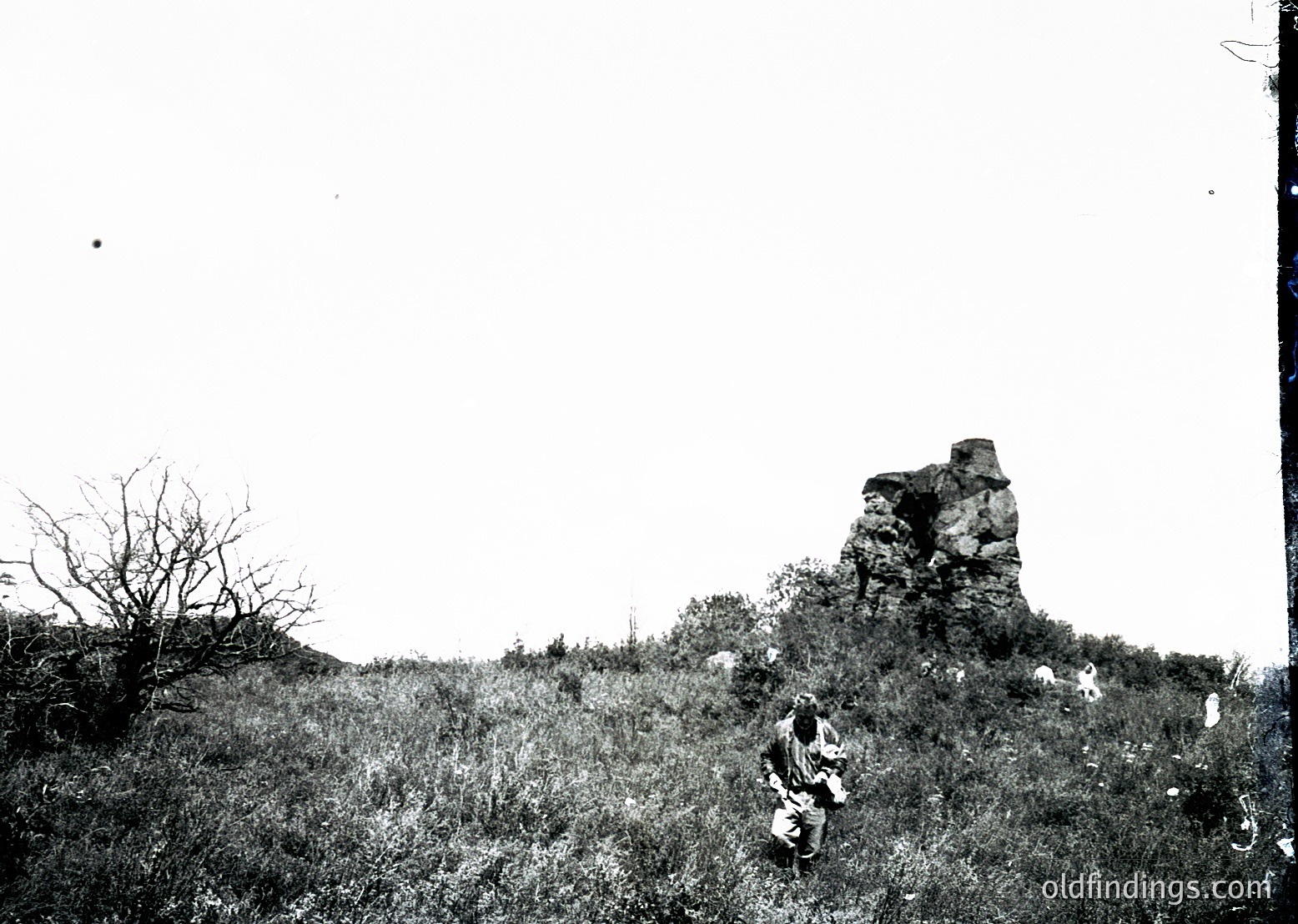Vintage black-and-white photo of a lone figure seated atop a grassy hillside, with a prominent rock formation in the background. The individual wears a long coat and hat, suggesting early-to-mid 20th-century attire. The rugged terrain and solitary pose evoke a sense of isolation and contemplation.