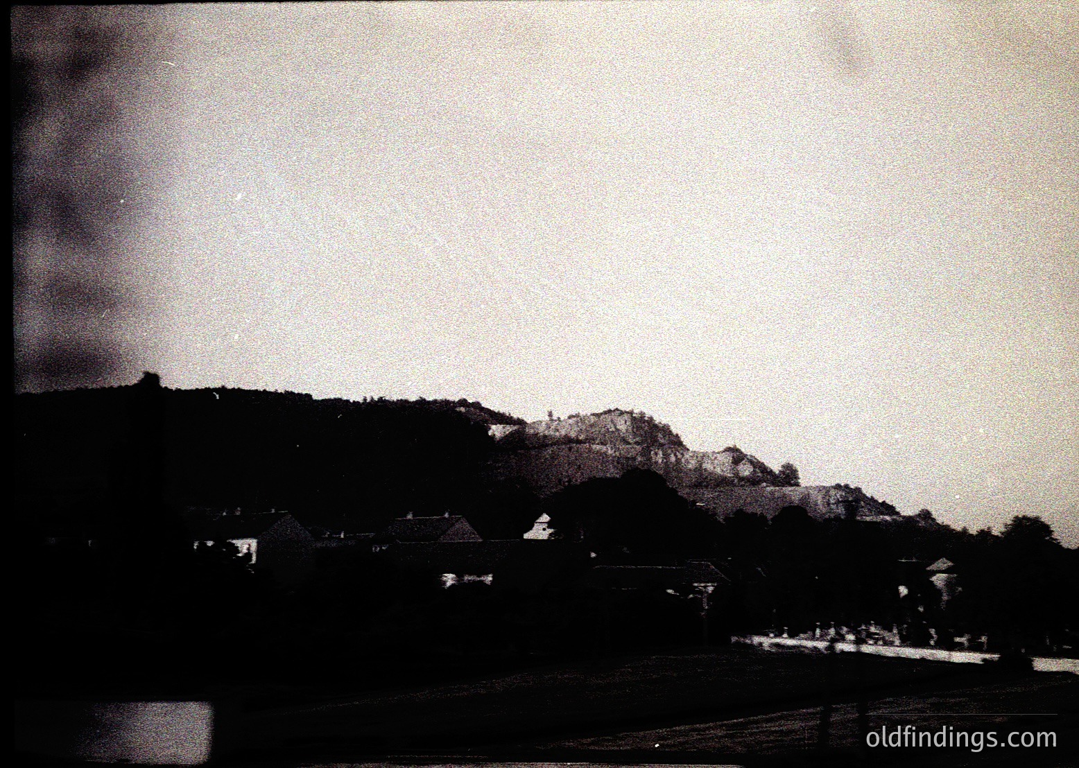 Vintage black-and-white landscape featuring a low-contrast rural scene with dense forest and rocky hillside. Small, clustered buildings suggest a village or hamlet. Faded film grain and slight overexposure hint at mid-20th century photography. Ideal for historical or nostalgic design references.