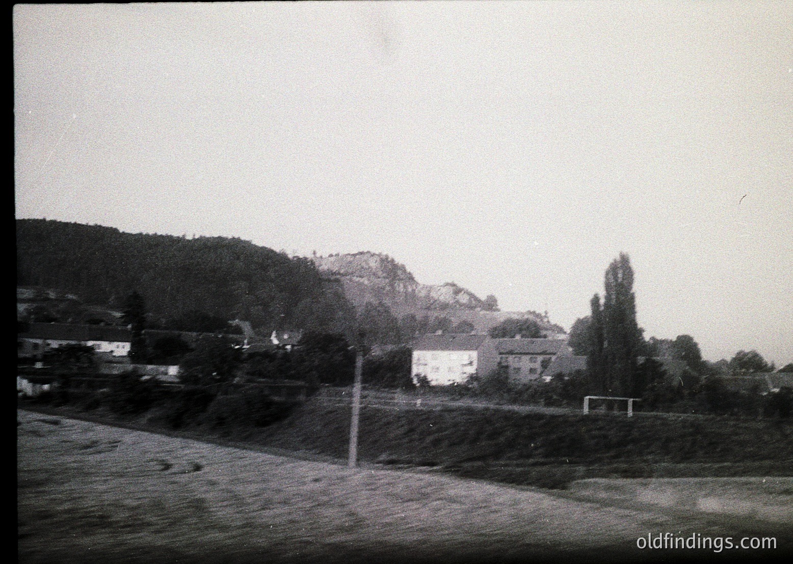 Vintage black-and-white rural landscape featuring a motion-blurred roadside view. Low-lying houses and dense greenery frame a soccer field with a goalpost. Hilly terrain and sparse trees dominate the background, suggesting a quiet, possibly European countryside. Likely mid-20th century.