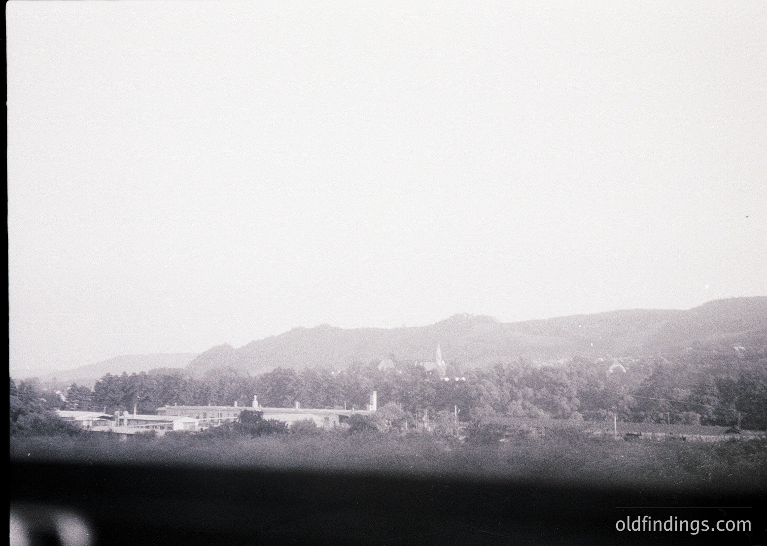 Vintage black-and-white landscape featuring a low-angle view of a large, symmetrical institutional building surrounded by dense forest. Distinctive spires and flat rooftops suggest a mid-20th-century architectural style. Rolling hills and misty horizon enhance the serene, isolated atmosphere.