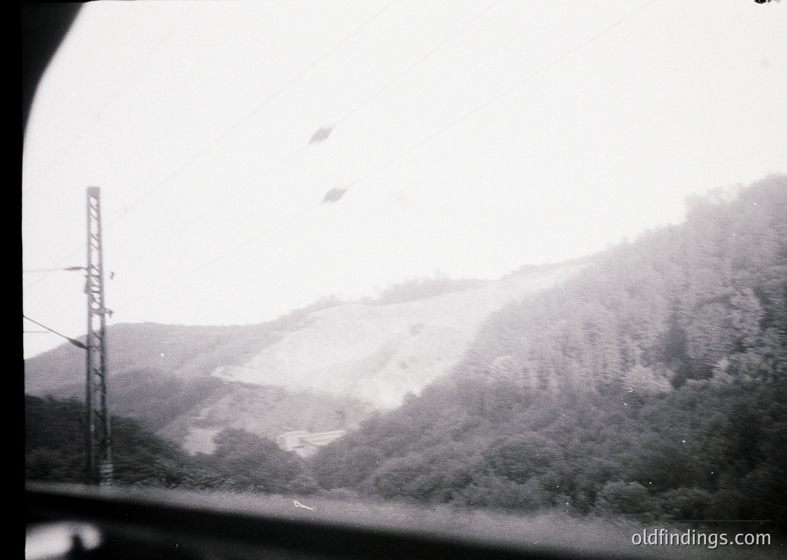 Black-and-white train window view of misty, snow-capped alpine ridge. Power lines and forest below suggest mid-20th century European rail travel. Fog obscures distant peaks, emphasizing atmospheric depth.
