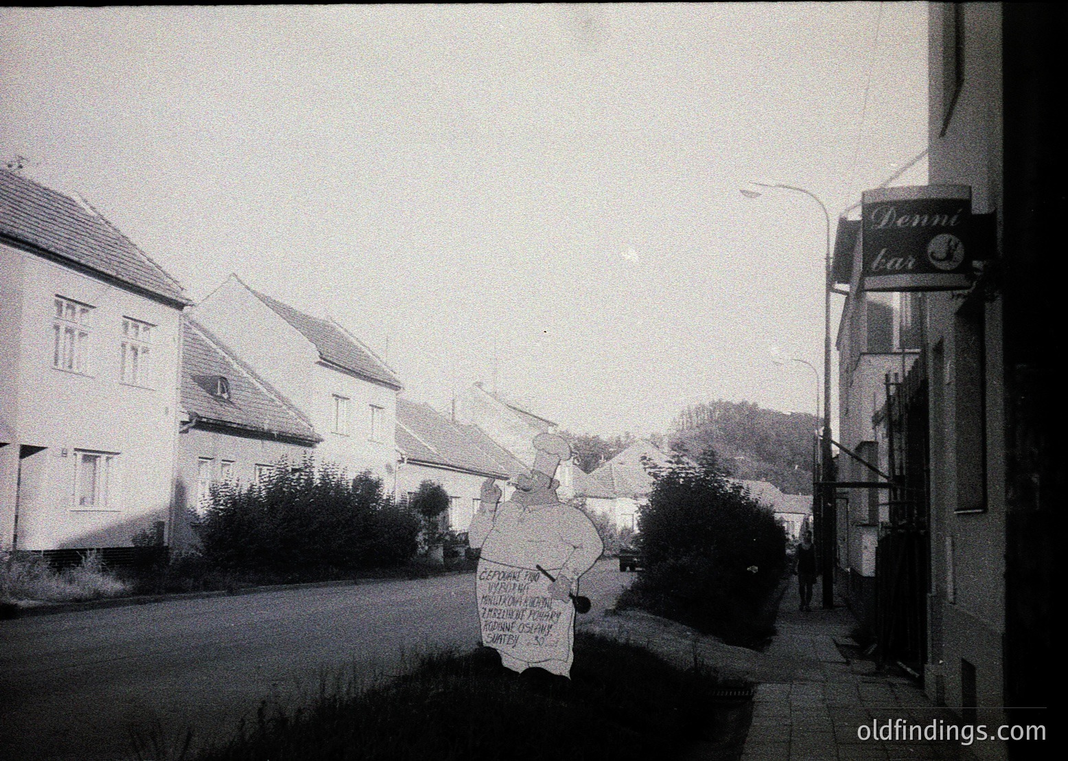 Vintage black-and-white street scene featuring a hand-painted protest sign in Cyrillic script, likely Bulgarian, with a "Denna bar" (Danube Bar) sign in the background. Residential buildings with pitched roofs and small gardens line the quiet road.