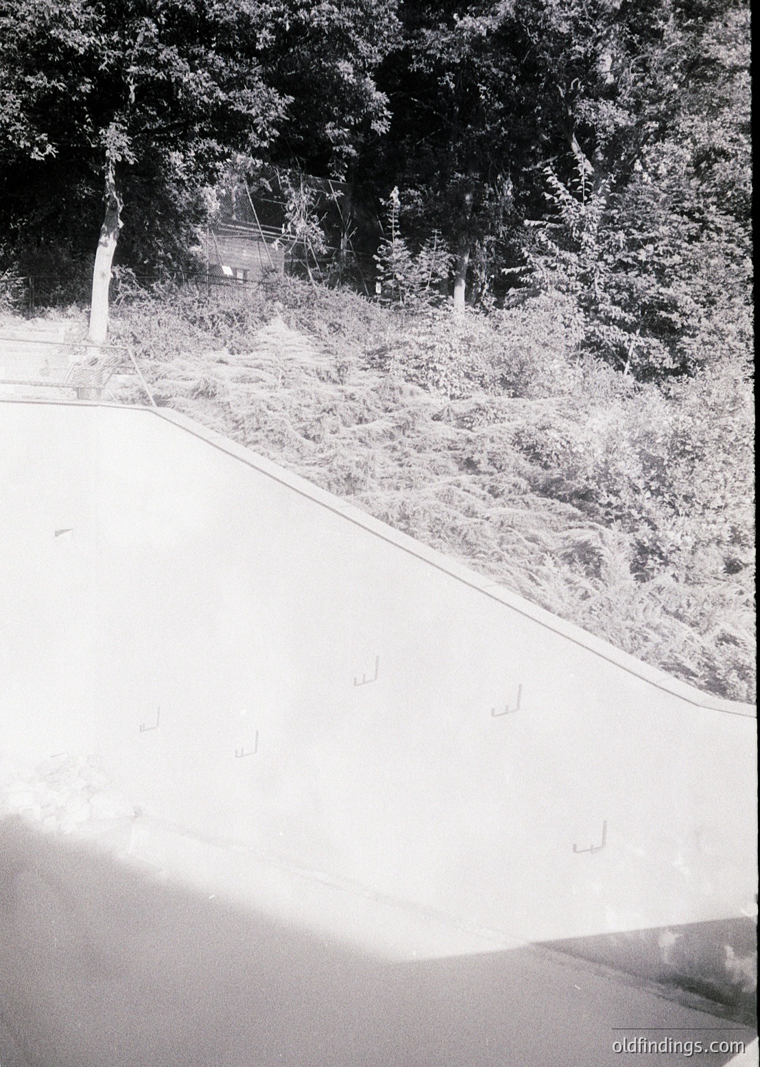 Vintage black-and-white shot of a winding rural road bordered by dense forest. Snow blankets the ground, with tire tracks visible. A small wooden bridge or gate peeks through the trees, suggesting a rural or forestry access point. Likely mid-20th century, possibly for historical or nostalgic design references.