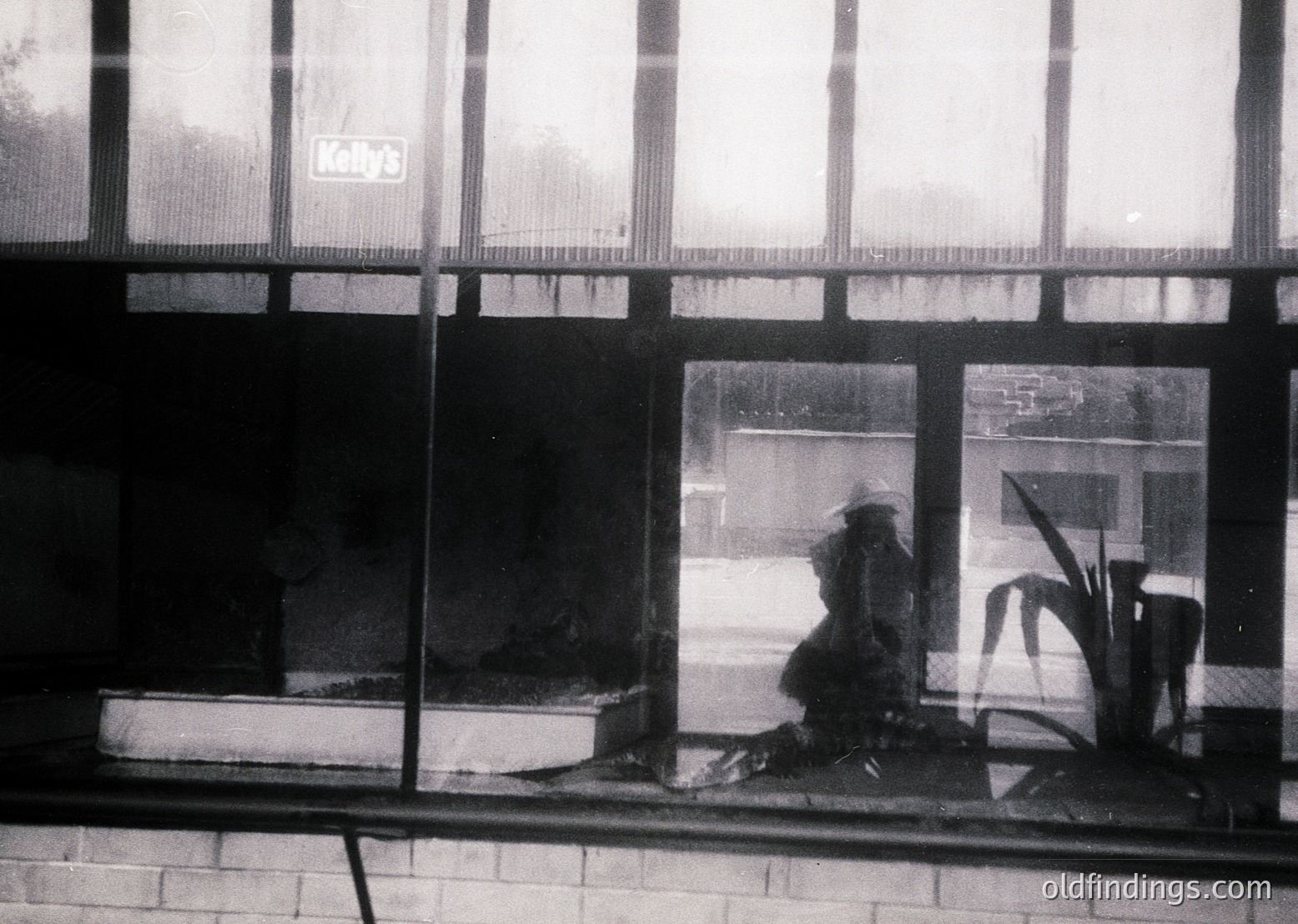 Black-and-white street scene captured through a storefront window, featuring a seated figure in a wide-brimmed hat and dark coat. Reflections reveal a quiet urban street with parked cars and distant buildings. Graffiti-like text ("Koby's") on the window frame suggests a commercial area. Mid-20th century (likely 1950s–1960s) urban architecture.