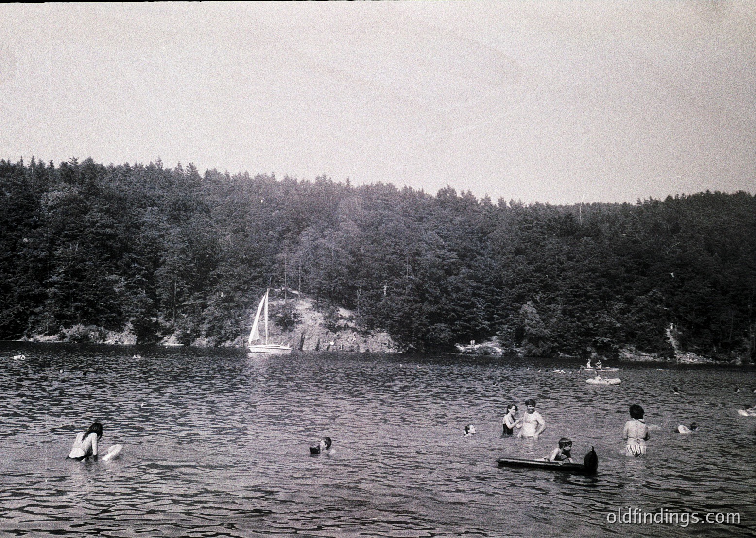 Vintage black-and-white seaside scene with 1950s-60s swimwear, including men in trunks and women in one-piece suits. Group of 8+ people swimming and wading in calm waters near rocky shore. Dense forest and sailboat in background.