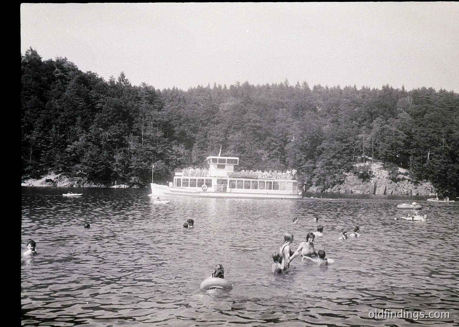 Vintage black-and-white photo of a mid-20th century lakeside scene. A group of people swim in calm waters near a docked ferryboat with "S.S. *name*" visible. Lush forested hills frame the scene, suggesting a recreational or tourist destination.
