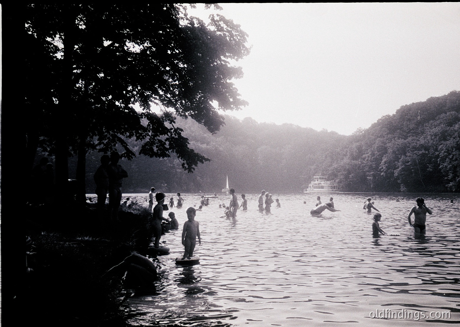 Black-and-white scene of a lakeside gathering, likely mid-20th century. Dense tree canopy frames a shallow lake where children and adults wade, swim, and play. A distant paddleboat and lush forest backdrop suggest a rural or park setting. Sunlight filters through leaves, casting soft shadows.