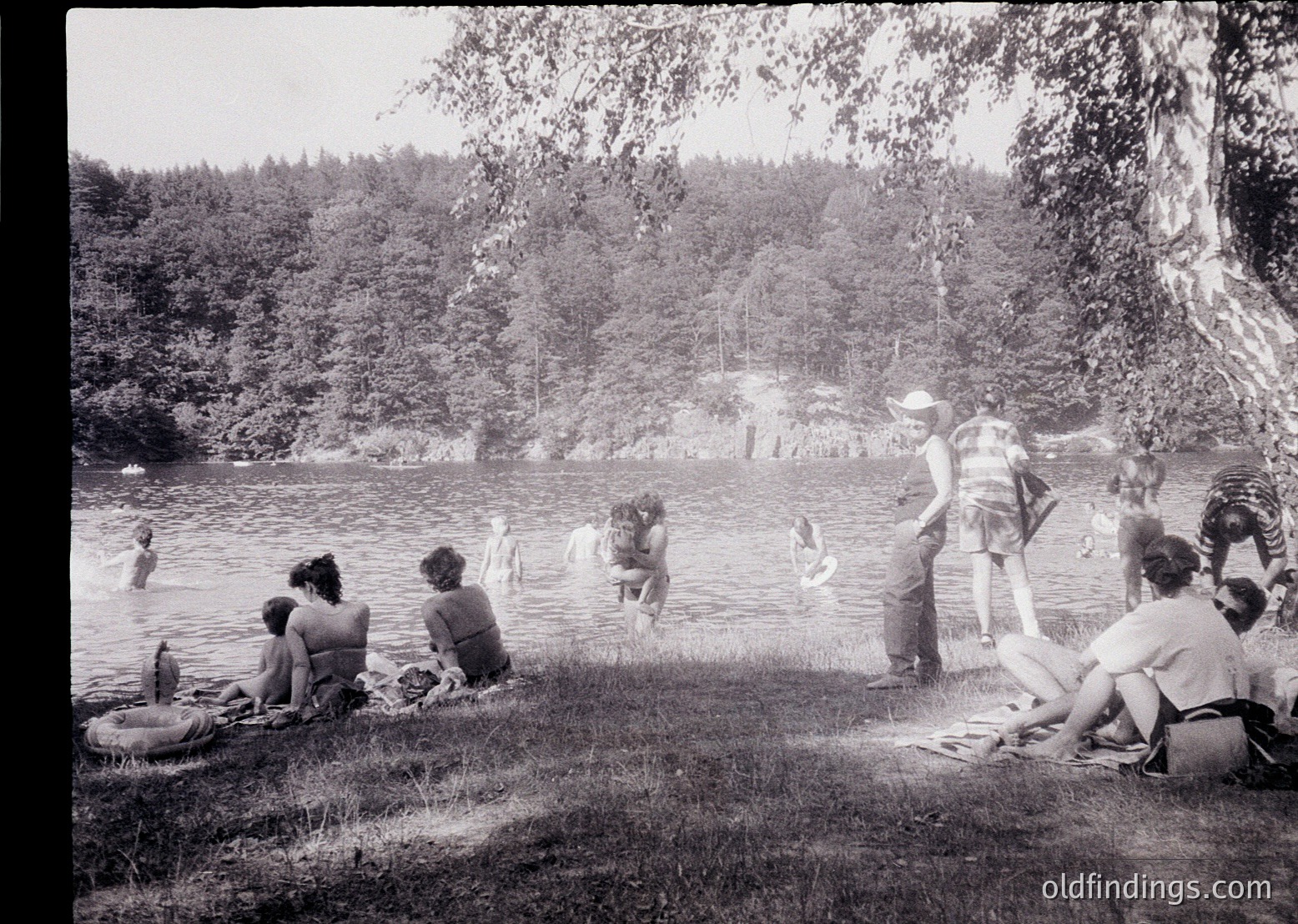Mid-20th century lakeside gathering with 1950s–60s swimwear and vintage floatation devices. Group of men and women lounging on grass near shallow water, others wading or swimming. Dense forest backdrop suggests rural or park setting. Black-and-white film captures candid, leisurely atmosphere.