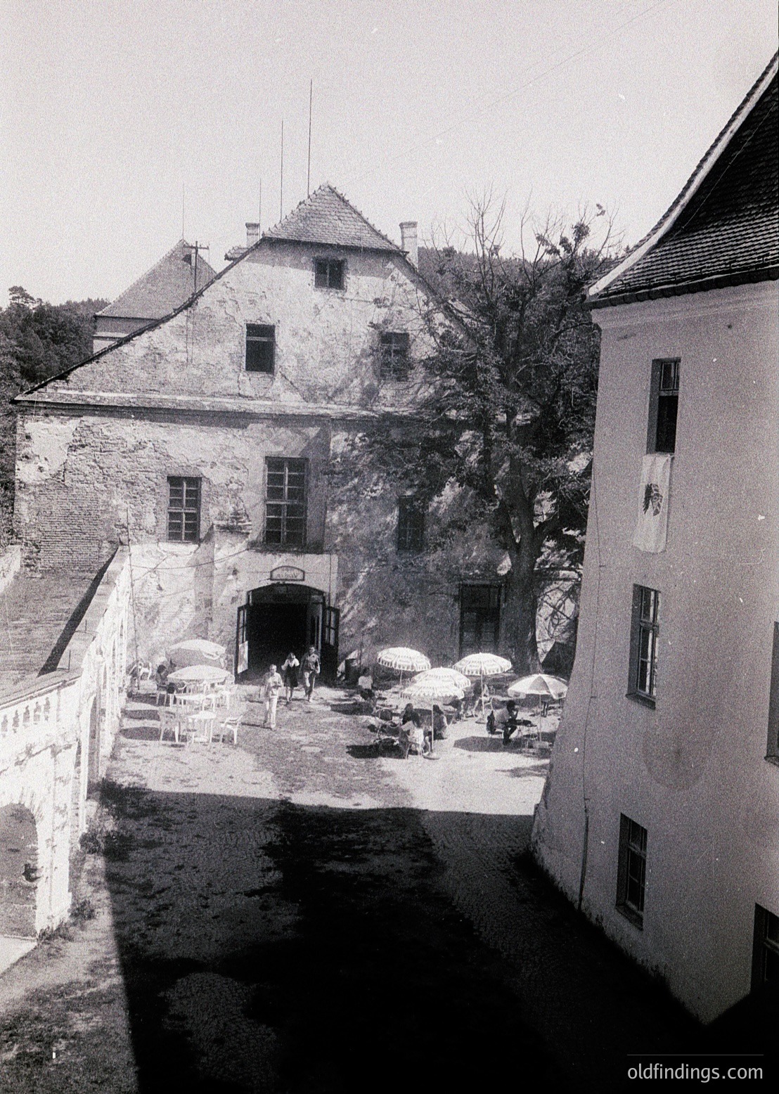 Historic stone courtyard with gabled building featuring arched entrance and small windows. Umbrellas and outdoor seating suggest a café or public gathering space. Likely Eastern European architecture, mid-20th century.