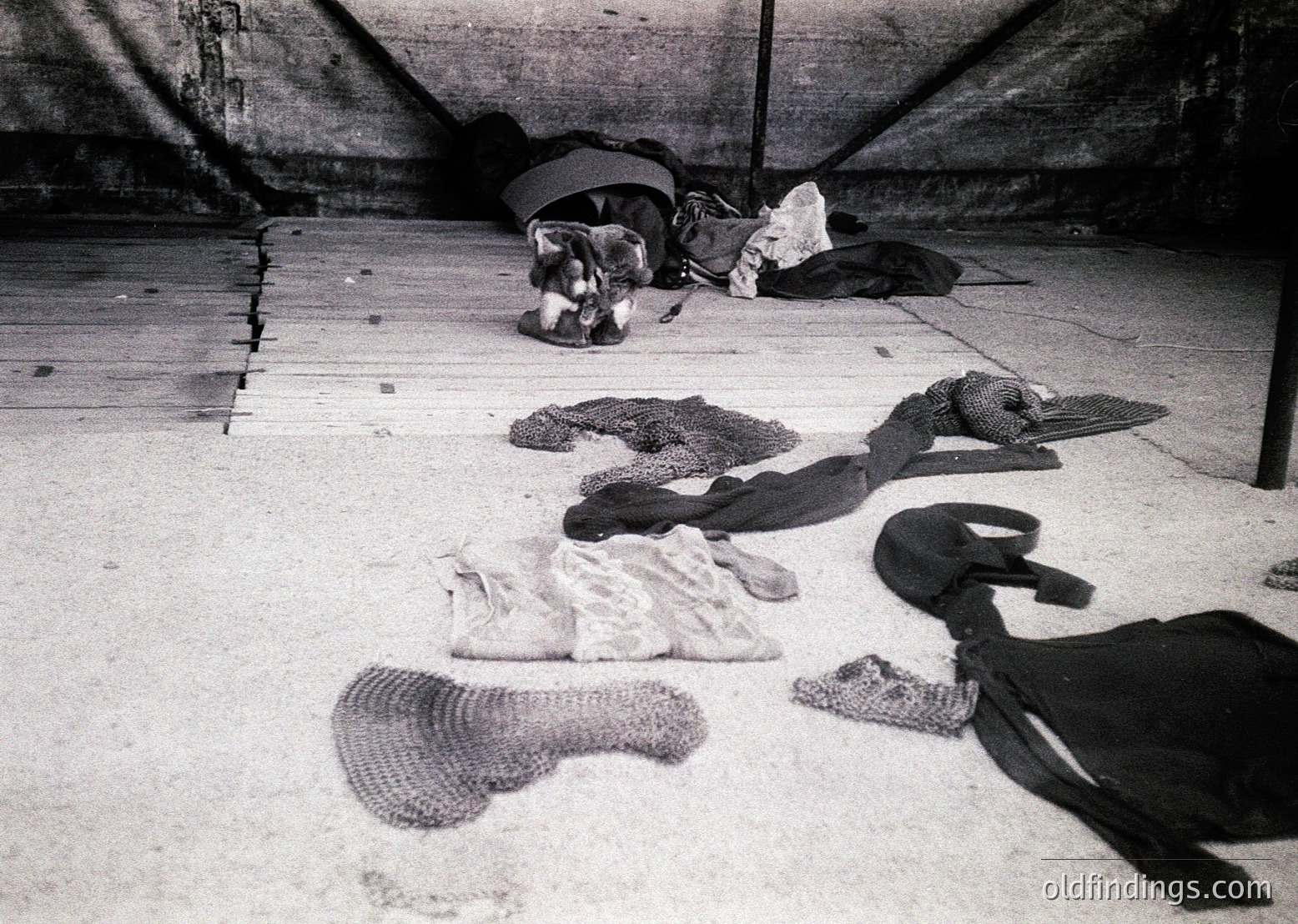 Black-and-white photo of discarded clothing and textiles on a concrete floor, likely a basement or storage area. Items include a knitted blanket, a sweater, a scarf, and a plastic bag. The scene suggests abandonment or post-use discard, possibly mid-20th century based on style and grain.