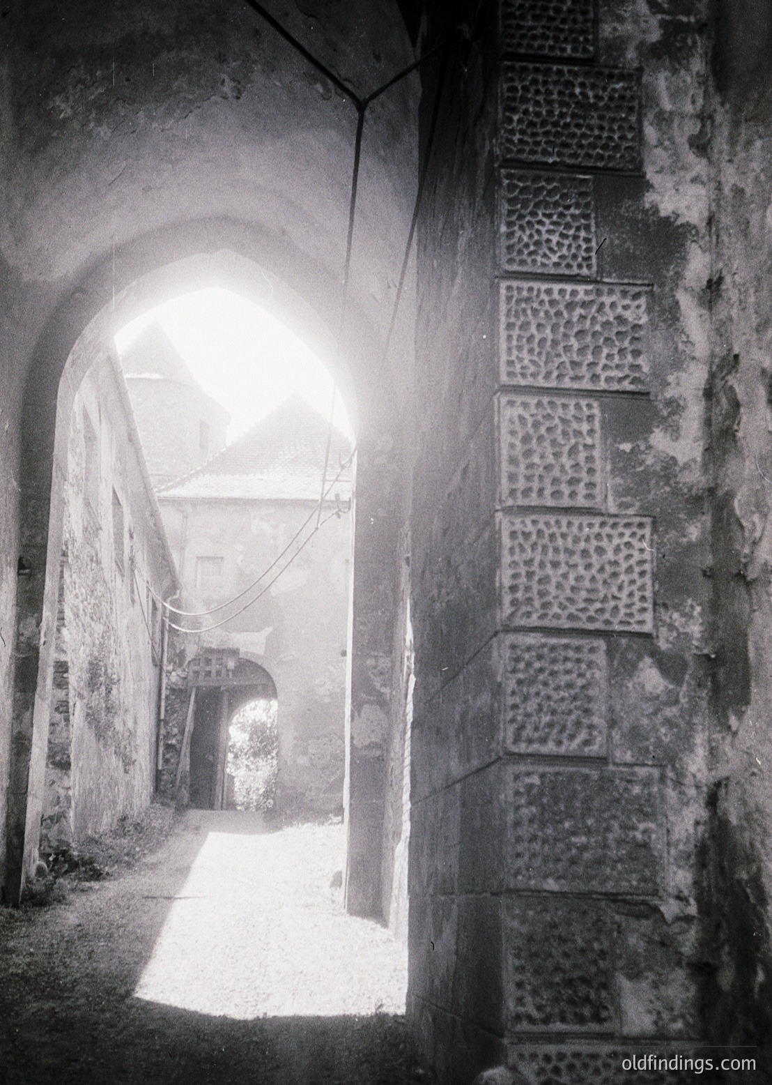 Sunlight filters through an arched stone passageway, illuminating a narrow alleyway with textured brick walls and cobblestone ground. The architectural details suggest historic European design, likely 19th–early 20th century.