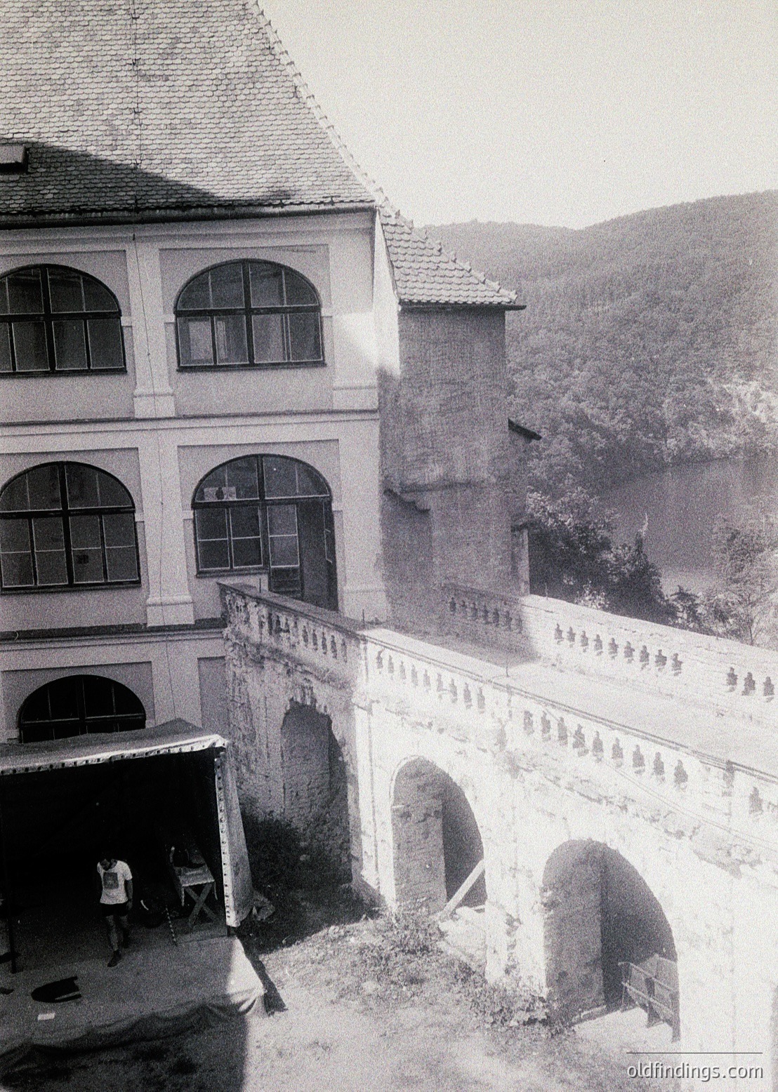 Neoclassical building with arched windows and arched passageways, likely a historic estate or monastery. Two figures in courtyard, one seated at a table. Mountainous backdrop suggests alpine or hilly region. Black-and-white, vintage aesthetic ().