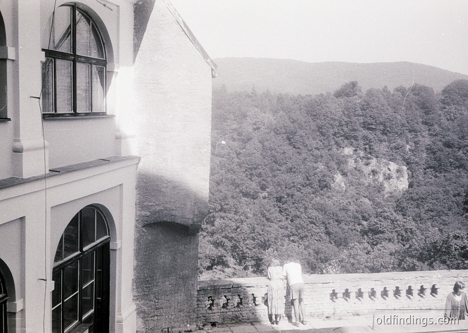 Mid-20th century black-and-white shot of a stone balcony overlooking lush, terraced hills. Three figures in vintage clothing pose near a carved stone railing with relief carvings. Adjacent building features arched windows and a curved facade. Likely European, possibly