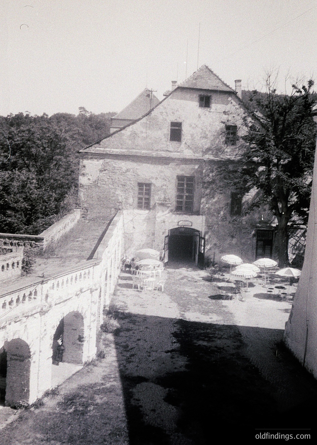 Historic stone courtyard with arched bridge leading to a two-story building featuring gabled roof and symmetrical windows. Umbrellas and outdoor seating suggest a café or public gathering space. Architectural style hints at 19th-century European influence.