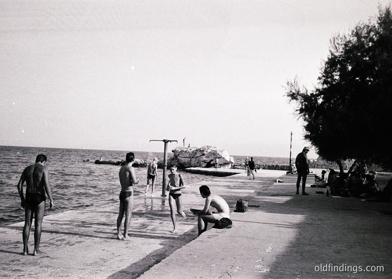 Black-and-white seaside scene with young men in swim trunks (1950s–60s style) by a concrete pier. One man sits reading, others stand or wade in shallow water near a small boat. Lifeguard tower and rocky breakwater visible.