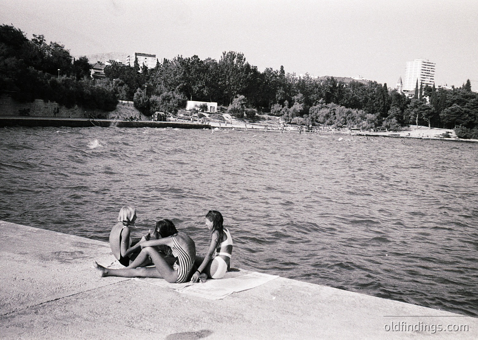 Three women in 1960s-style swimsuits sit on a concrete ledge by a lake, backs to camera. Mid-century urban park in background with trees, low buildings, and distant high-rise. Classic black-and-white composition.
