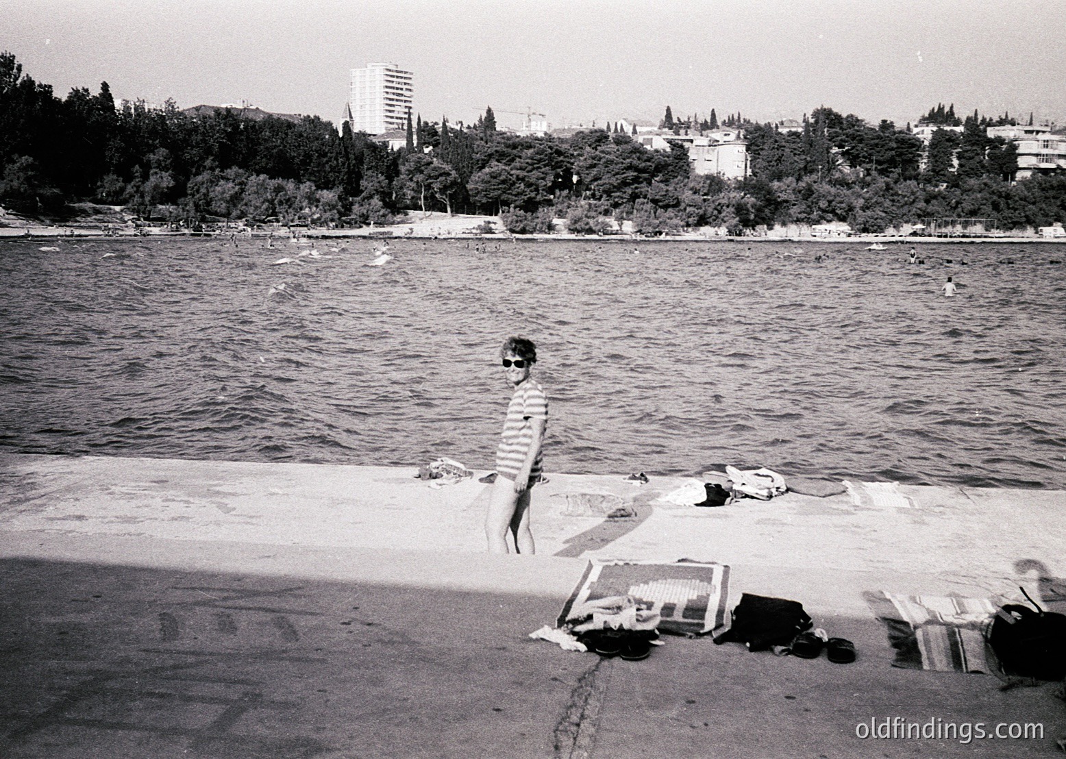 Mid-20th century seaside scene: a lone child in shorts stands on a sandy shore, framed by scattered towels and a rolled blanket. Urban skyline with high-rise and trees in background suggests a coastal city. Black-and-white grain captures 1950s–1960s era.