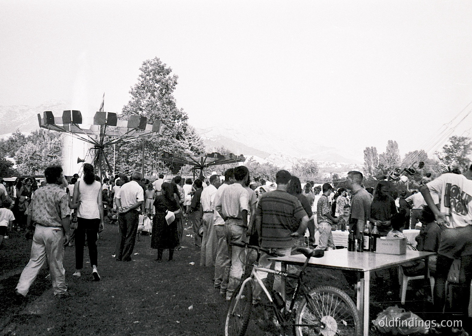 Crowded outdoor gathering in a rural setting, likely 1970s–1980s Eastern Europe. Central structure features a tall, multi-sided flagpole with a red star emblem, surrounded by people in casual attire. Tables with white tablecloths and bicycles suggest a communal event or festival. Hilly terrain and trees in background.