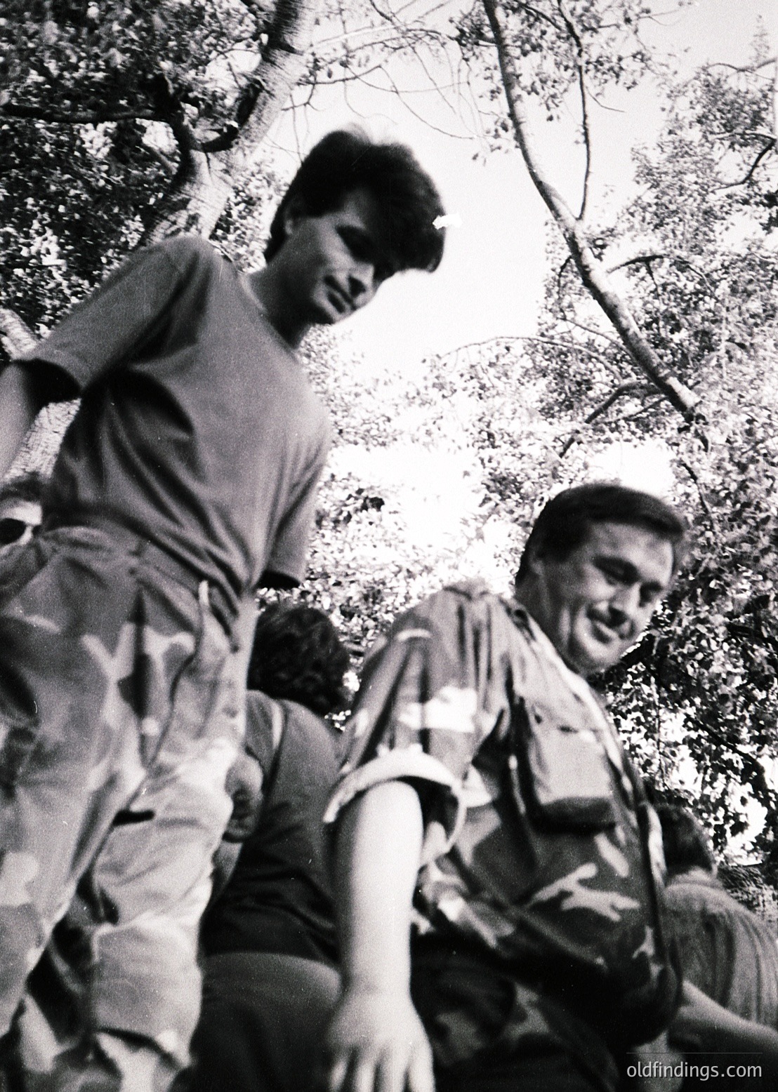 Black-and-white candid of two men laughing while hiking, framed by dense forest foliage. Mid-20th century outdoor attire suggests or . Casual, joyful group activity in nature.
