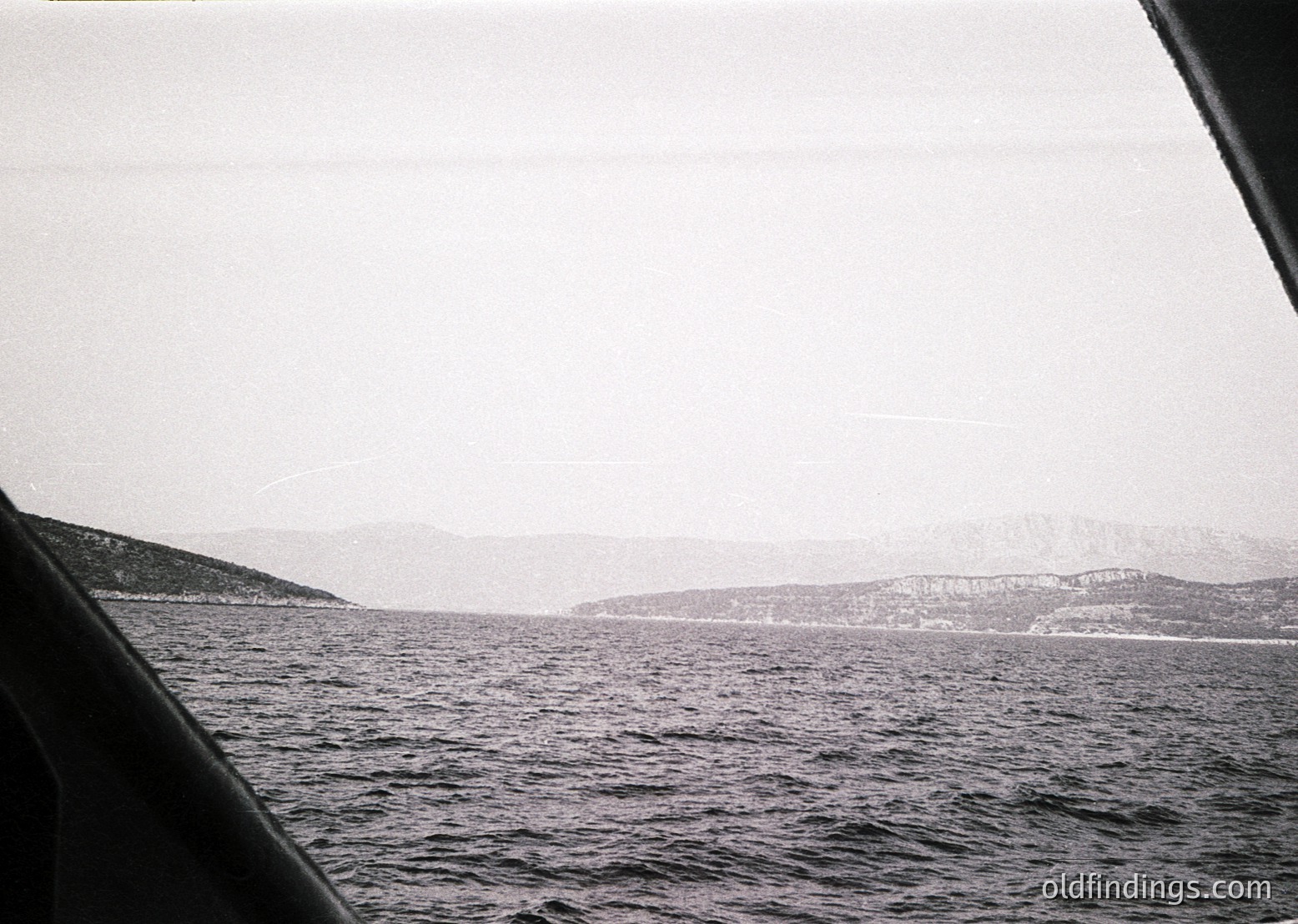 Black-and-white seascape shot from a boat’s perspective, showing choppy waters and distant landmass with faint outlines of trees. Composition emphasizes horizon and misty atmosphere, likely mid-20th century maritime photography. Ideal for vintage travel or nautical archives.