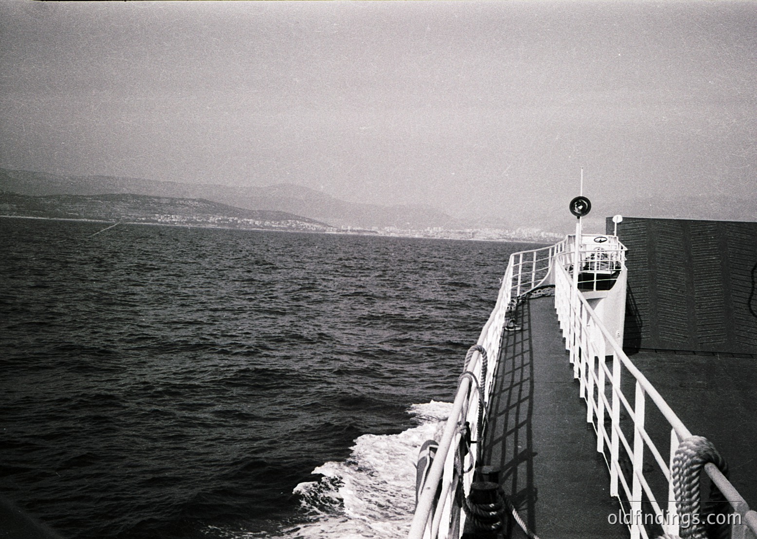 Black-and-white shot of a ferry’s bow cutting through calm waters, with distant coastal hills and misty horizon. Mid-20th century maritime design visible in railings and hull. Evokes midcentury travel or industrial-era coastal transport.