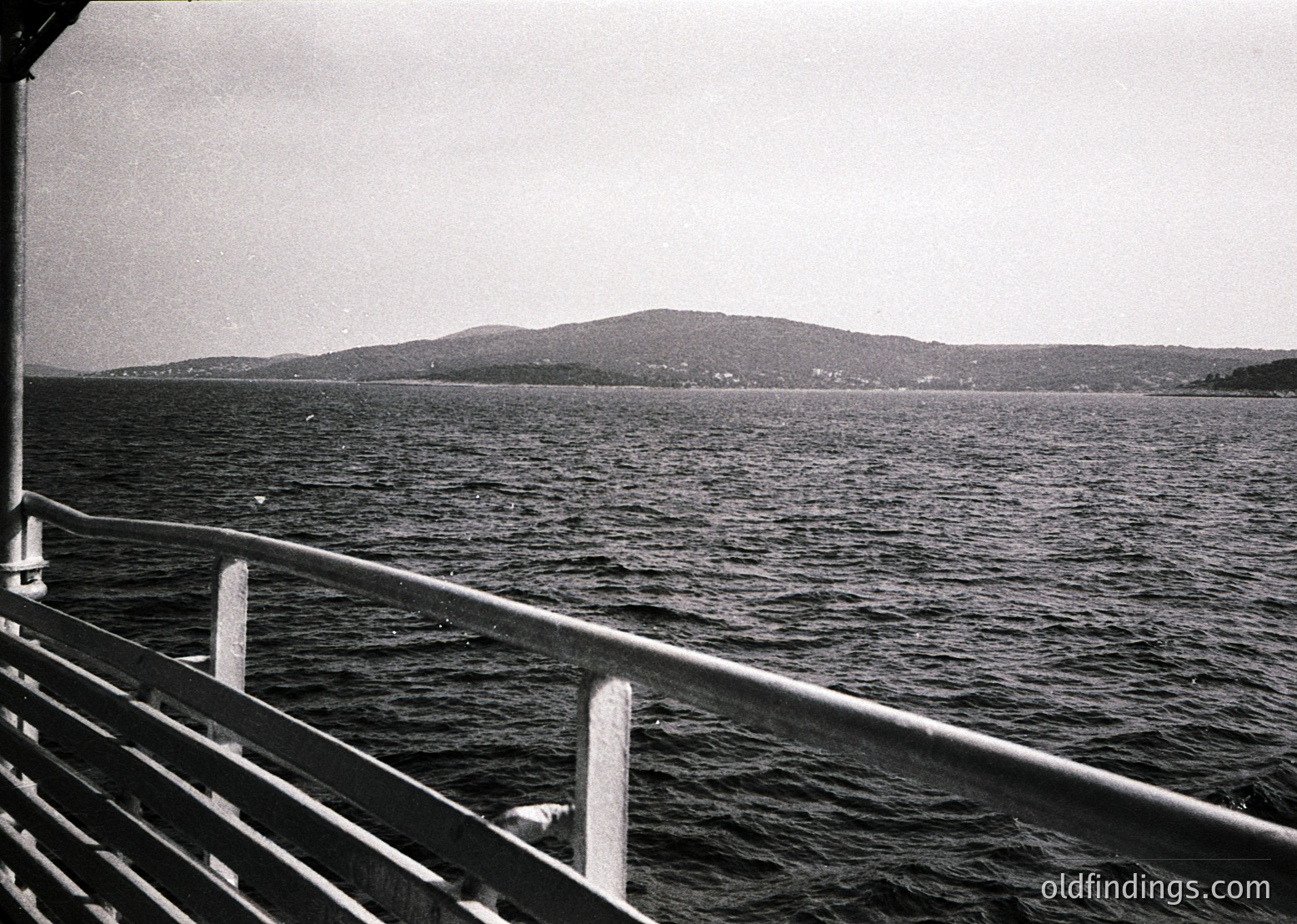 Black-and-white seascape shot from a ship’s railing, showing calm waters and distant hilly coastline. Mid-20th century maritime travel aesthetic with minimal human presence.