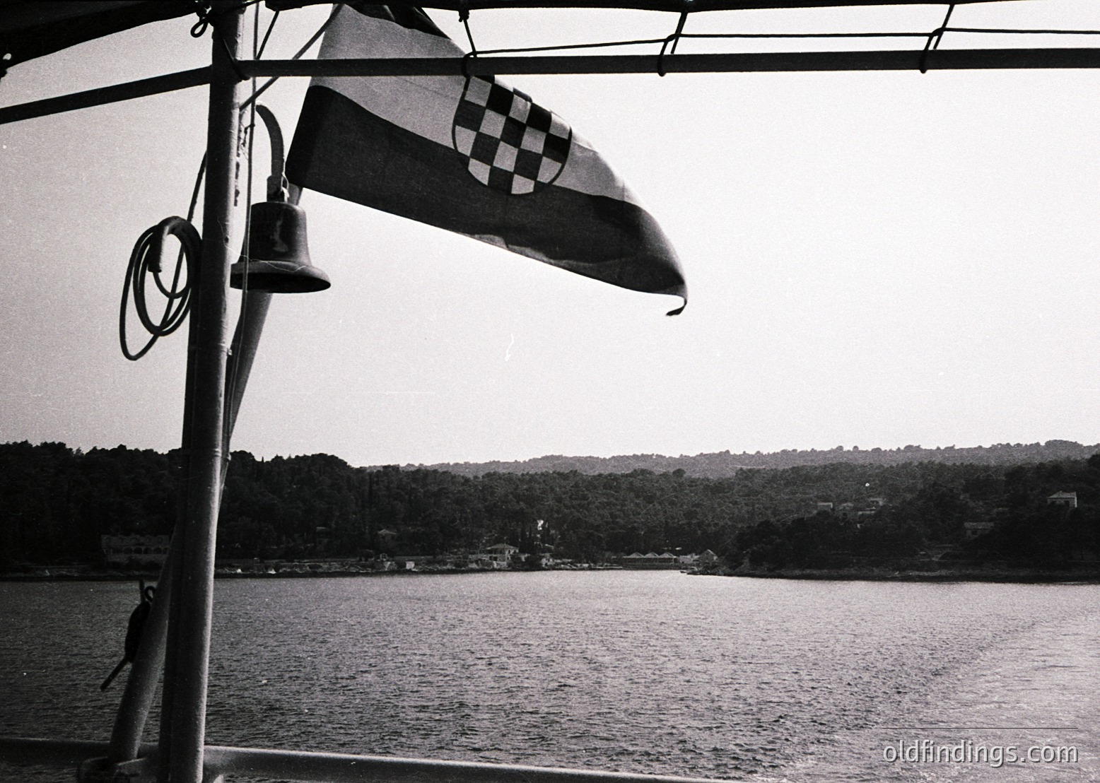 Croatian flag with checkerboard pattern waves over calm waters. Bell and rope detail on ship’s rail. Dense forested coastline in background. Black-and-white vintage maritime aesthetic. Likely Adriatic Sea region.