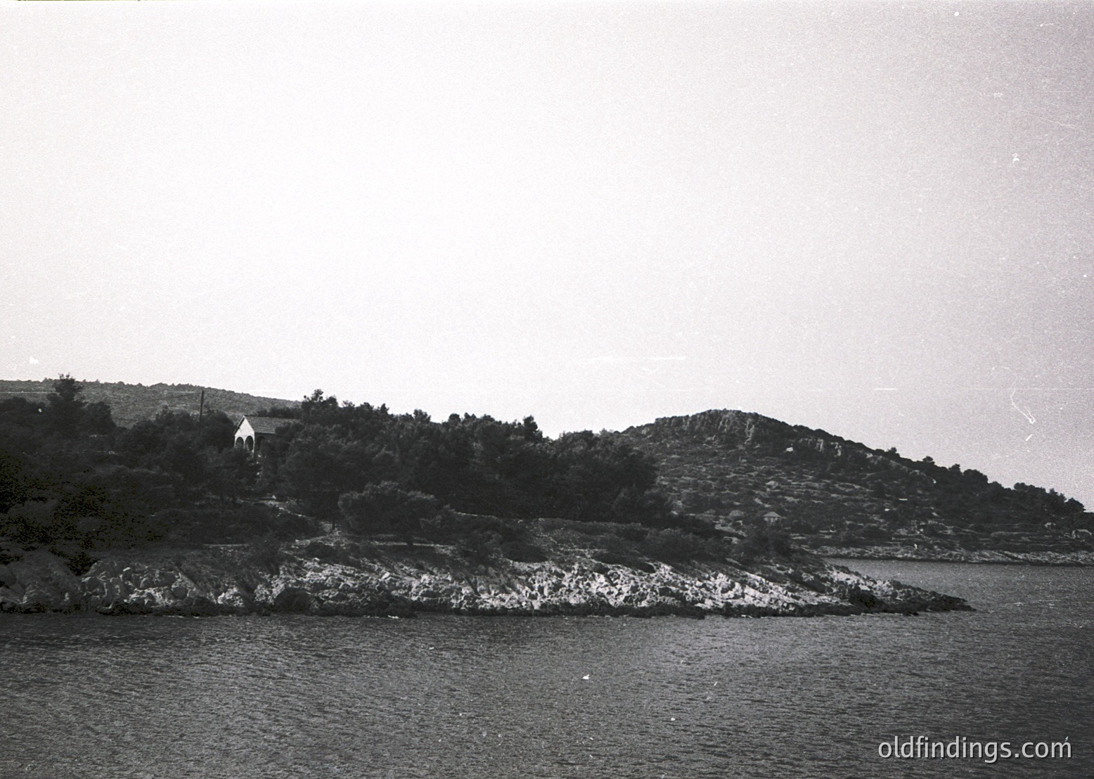 Black-and-white coastal scene featuring jagged rocky shoreline and calm waters. A lone figure stands atop a hillside covered in sparse vegetation, overlooking the sea. Mid-20th century aesthetic with high-contrast lighting.