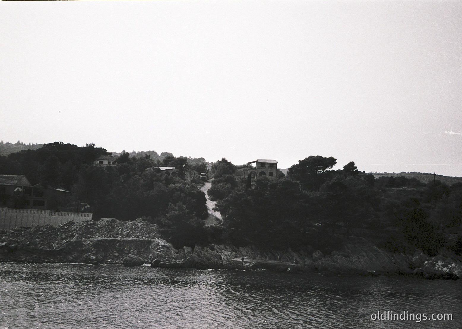 Black-and-white coastal landscape featuring a rocky shoreline and dense greenery. Mid-20th century architecture with flat-roofed buildings nestled among trees, likely Mediterranean or Balkan region. Waterfront path suggests historical or rural setting.
