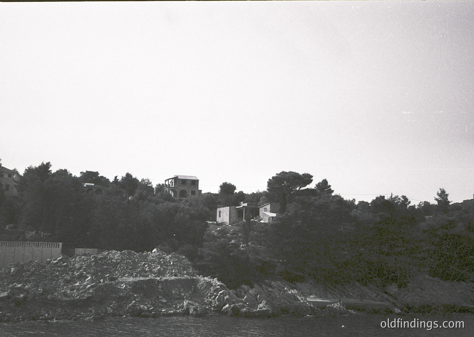 Black-and-white coastal landscape featuring two-story Mediterranean-style homes perched on rocky terrain. Dense greenery surrounds structures, with a rocky shoreline and water visible below. Likely Mediterranean region, mid-20th century.