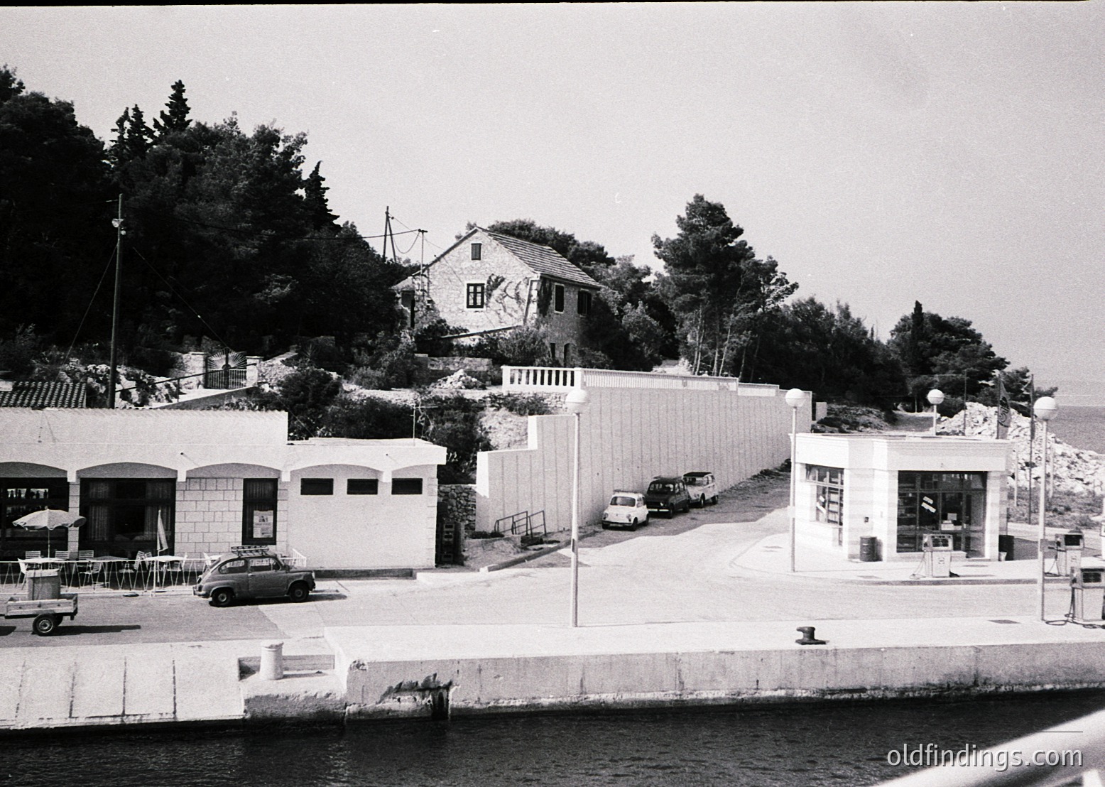 Mid-century seaside complex with Brutalist concrete structures, arched entryways, and minimalist design. Two-story building with graffiti on upper facade. Vintage cars parked along a paved promenade. Waterfront location with stone retaining wall. Likely 1960s–1970s coastal architecture.