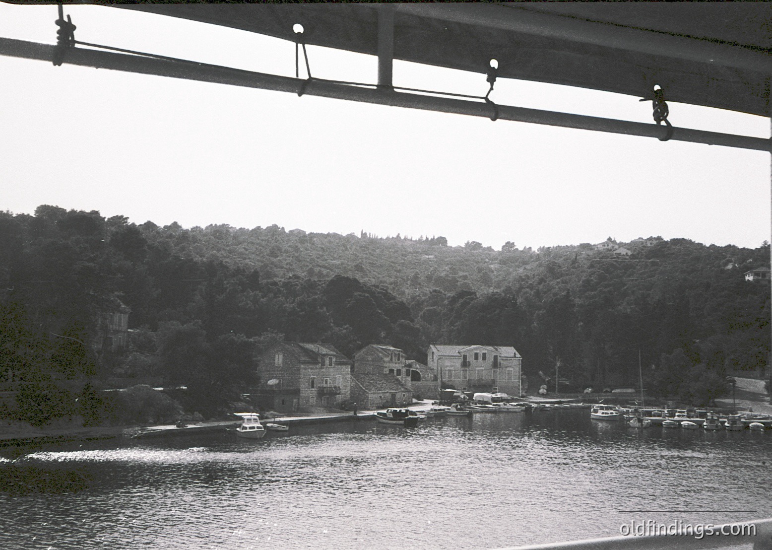 Black-and-white coastal scene featuring a bridge with hanging light fixtures framing the view. Below, a serene harbor with boats docked along a rocky shoreline, surrounded by dense, forested hills. Mid-20th century architecture visible in the background.