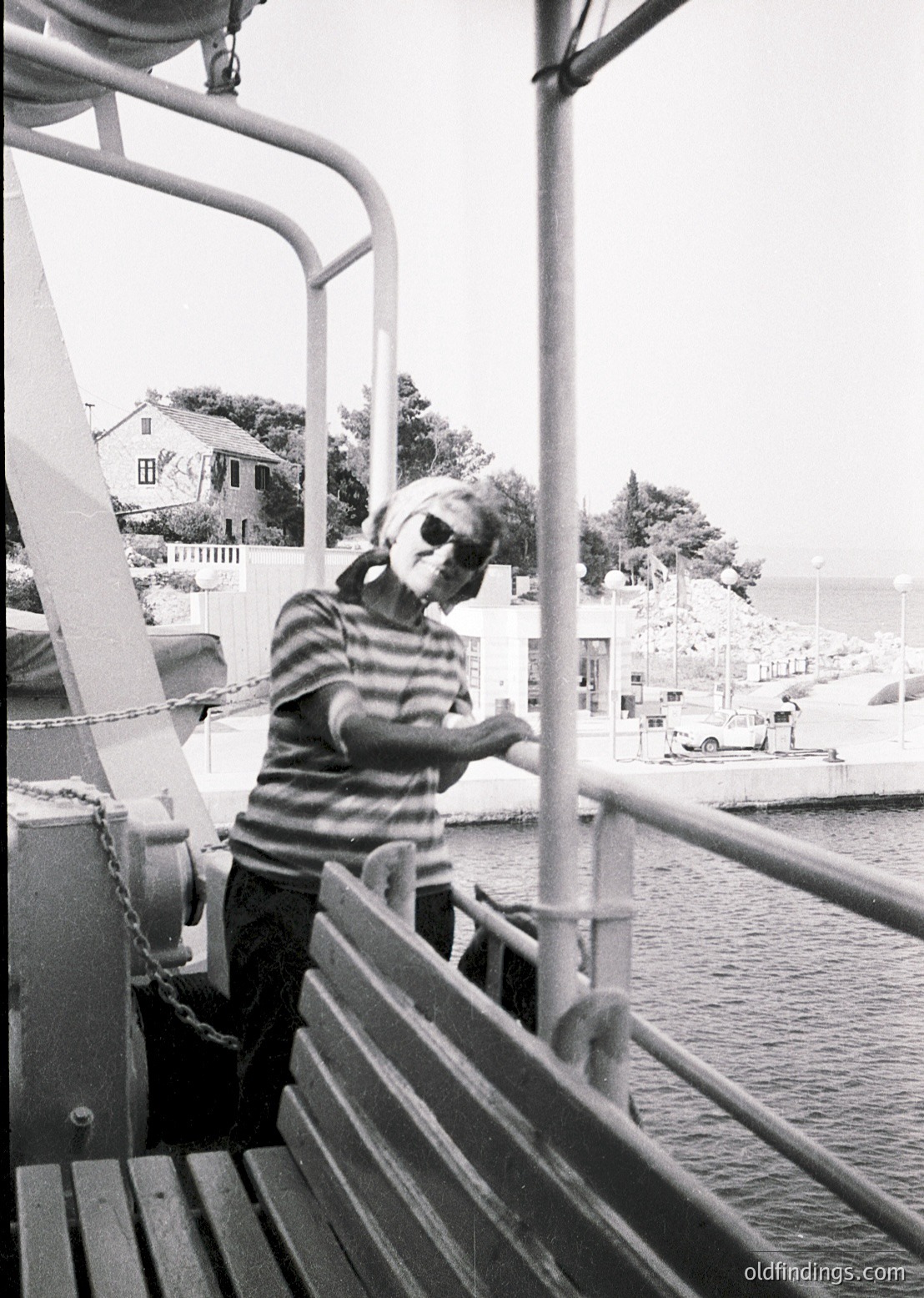 Black-and-white photo of a man in a striped sweater leaning on a ferry railing, overlooking a coastal town. Mid-20th century maritime setting with industrial railings and a distant dockside.