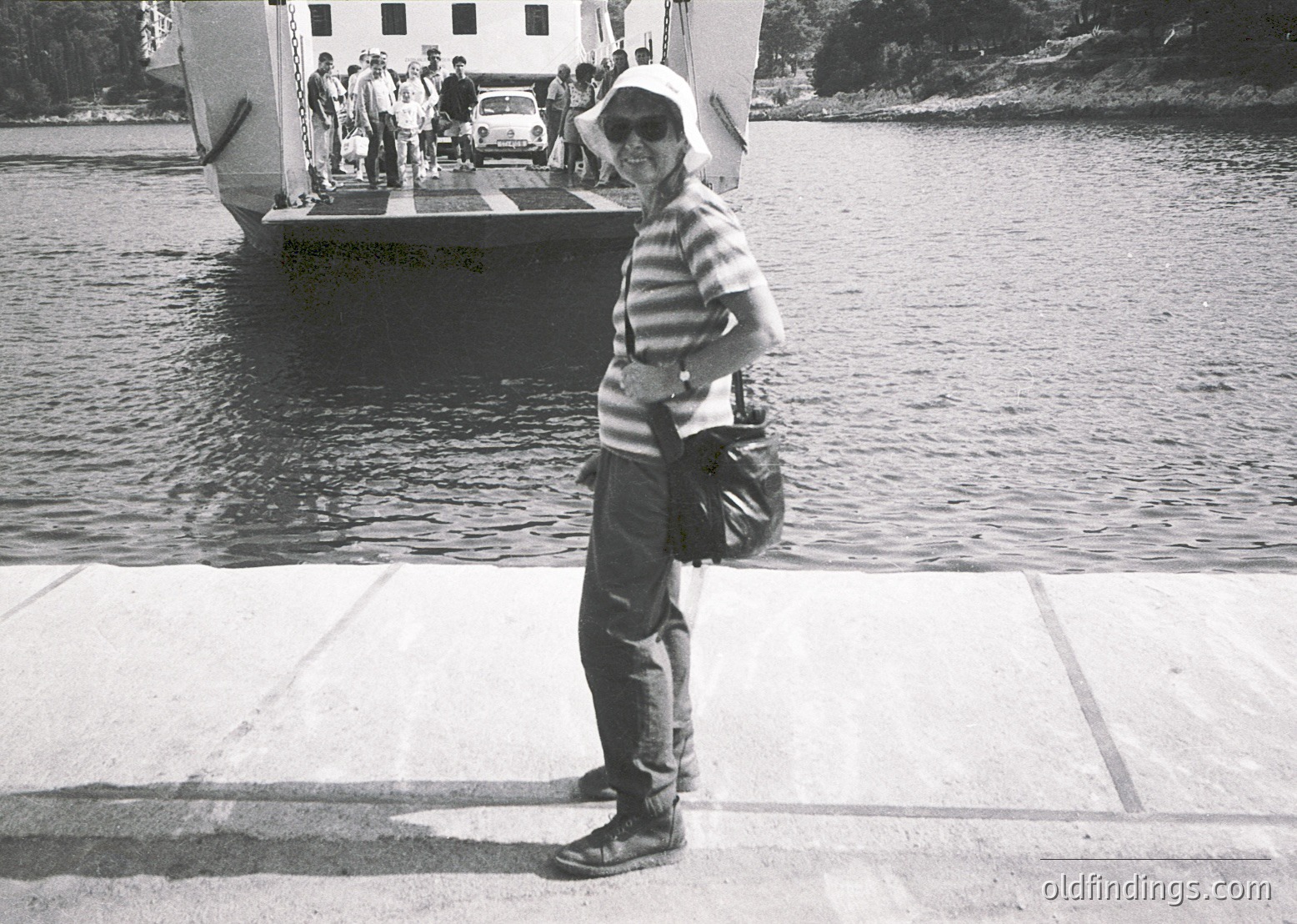 Mid-20th century ferry scene: Woman in striped blouse, wide-brim hat, and bag stands on concrete pier beside calm water. Classic car and vintage ferry in background with passengers boarding.