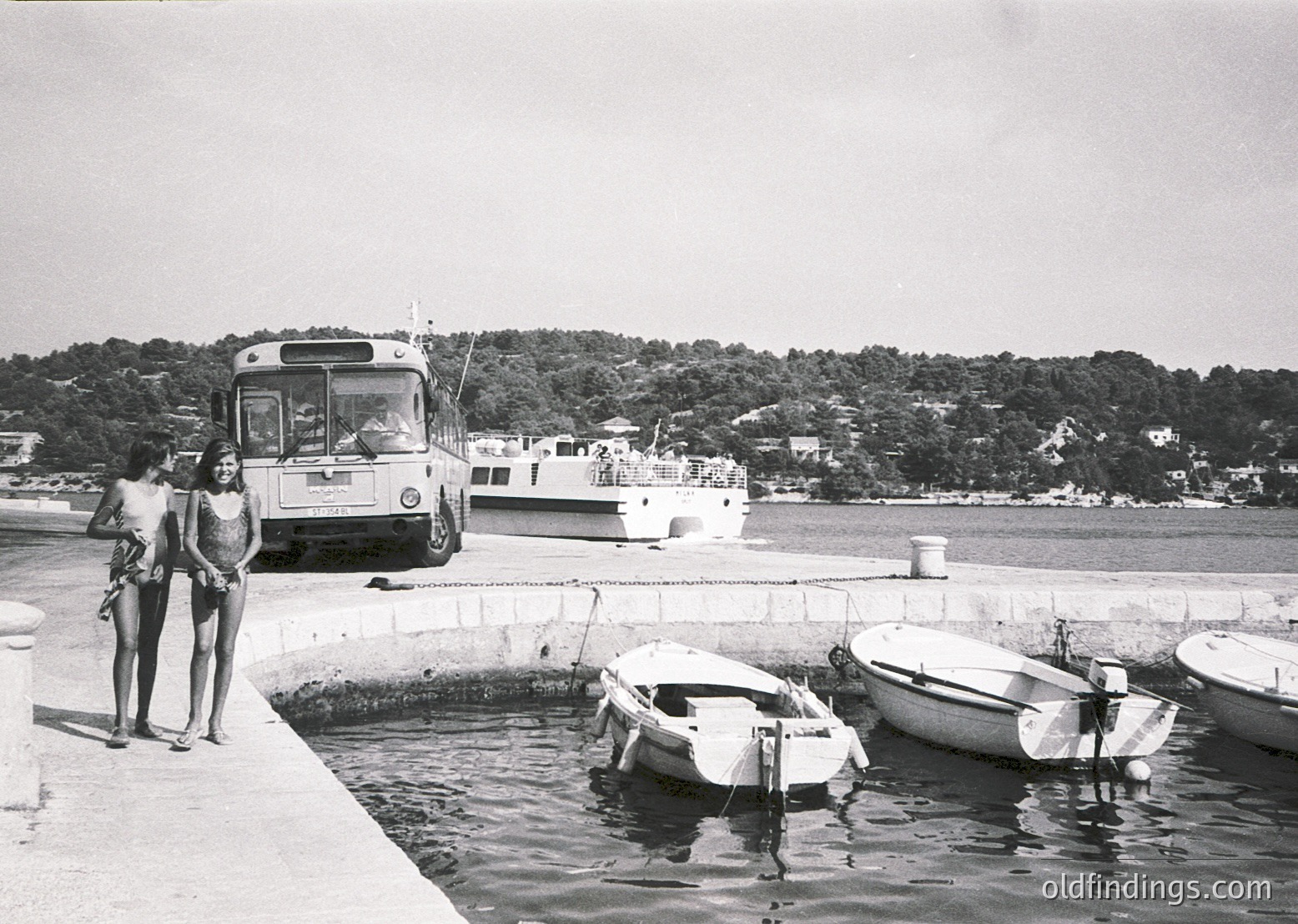 Two women in 1960s swimwear pose by a seaside pier, near a vintage ferry boat and small boats. Mid-century concrete harbor structures and lush green hills in background suggest a Mediterranean coastal town.