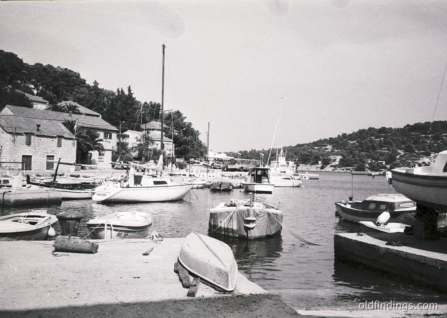 Black-and-white coastal marina with small boats docked along a concrete pier, surrounded by low-rise buildings with flat roofs. Wooden and fiberglass vessels, including sailboats and motorboats, line the waterfront. Dense greenery and hills frame the scene, suggesting a sheltered harbor. Likely Mediterranean or Adriatic region, mid-20th century.