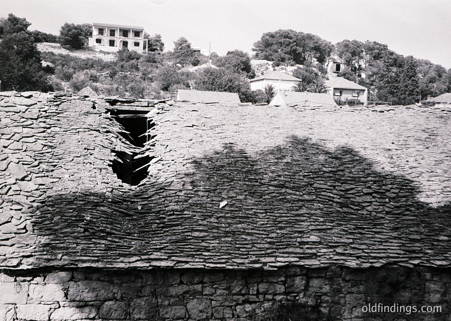 Ancient stone-walled ruins with a deep, vertical fissure in the foreground, likely a collapsed or eroded section. Midground shows scattered stone remnants and vegetation. Background features sparse, modest buildings and dense tree cover on a hillside. Black-and-white, suggesting mid-20th century archaeological or historical documentation.