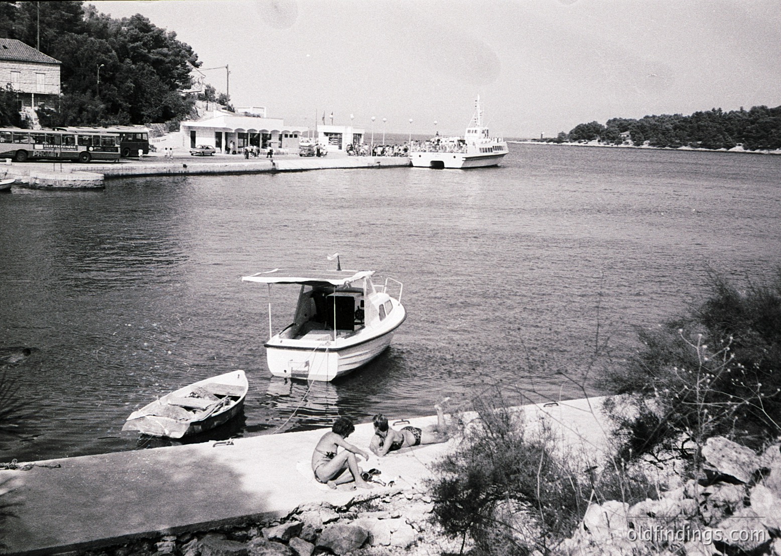 Mid-20th century seaside scene with a small motorboat docked near shore, two individuals sitting on rocky terrain. In background, a larger vessel and a building with a bus stop sign (, , , , ).