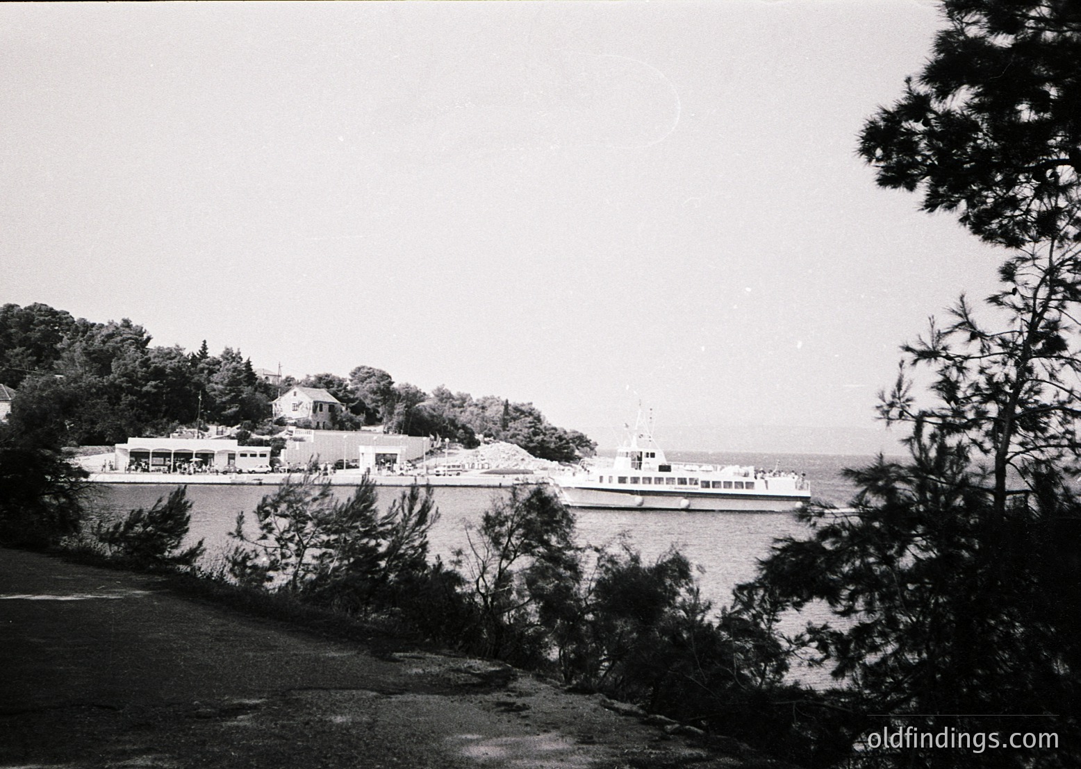 Black-and-white coastal scene featuring a mid-20th century ferry docked at a seaside promenade. Prominent low-rise buildings with flat roofs and balconies line the waterfront, likely a resort or port town. Dense greenery frames the left side, suggesting a forested or park area. The ferry’s white hull and red funnel indicate a Soviet-era vessel, possibly from the 1950s–1970s. The calm sea and overcast sky evoke a tranquil, utilitarian seaside atmosphere.