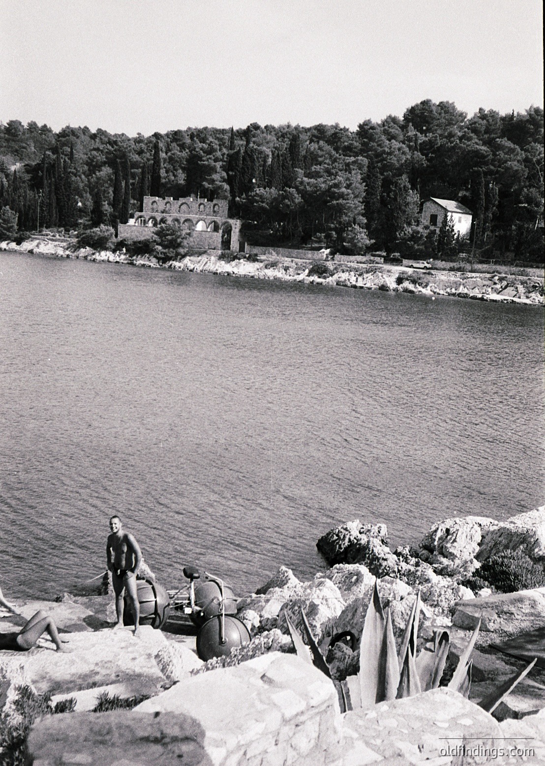 Black-and-white coastal scene featuring rugged rocky shore with two men in mid-20th century attire—one standing, one crouching—near a small boat. Linen or towels draped over rocks suggest a seaside retreat. Distant waterfront villa with arched windows and dense forested hillside in background. Likely Mediterranean, 1950s-60s.