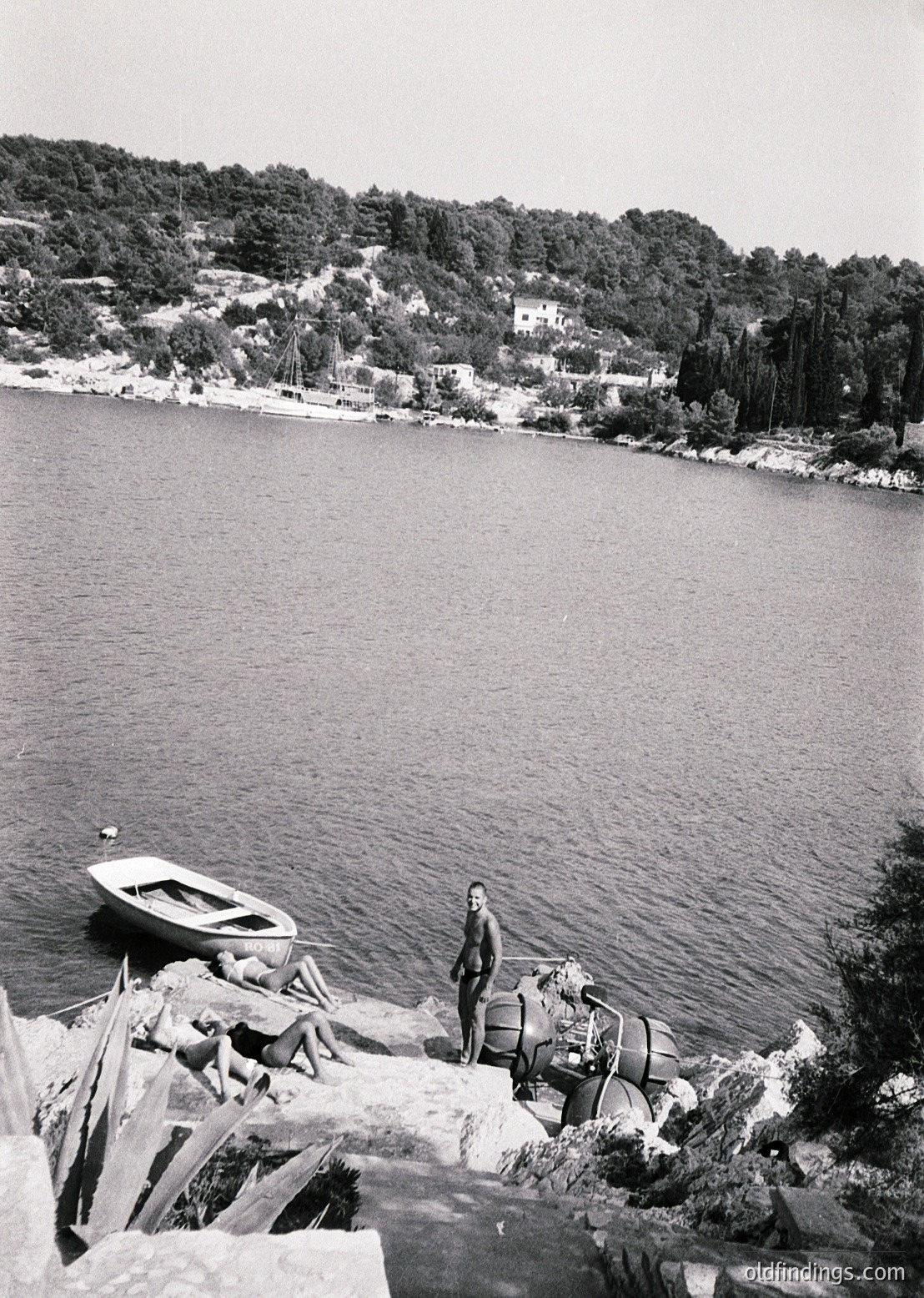 Black-and-white coastal scene featuring a man in mid-20th-century attire standing on rocky shore beside a small wooden boat. Lifebuoy and fishing gear visible. Dense forested hillside and scattered homes in background. Likely Mediterranean or Adriatic region, 1950s-1960s.