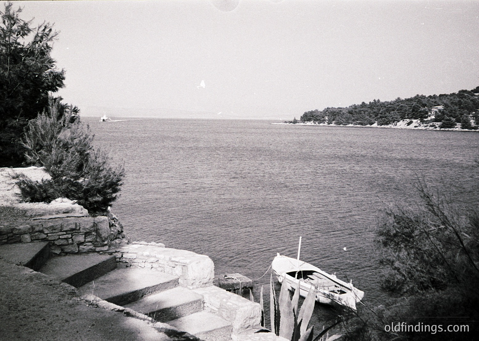 Black-and-white coastal scene featuring a stone staircase descending to a weathered wooden boat docked near calm waters. Dense tree line frames the right horizon, while a lone sailboat glides in the distance. Mid-20th century seaside architecture hints at a quiet, possibly Mediterranean or Eastern European lakeside retreat.