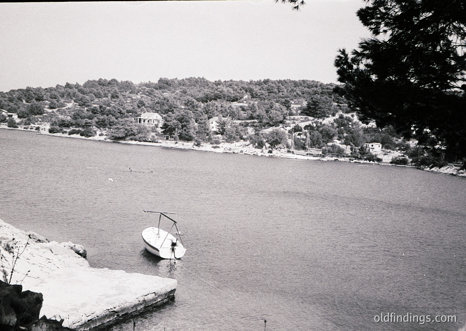 Black-and-white coastal scene featuring a small sailboat anchored near rocky shore. Dense forested hillside with scattered buildings and pathways in background. Mid-20th century seaside architecture visible. Calm water reflects muted light.